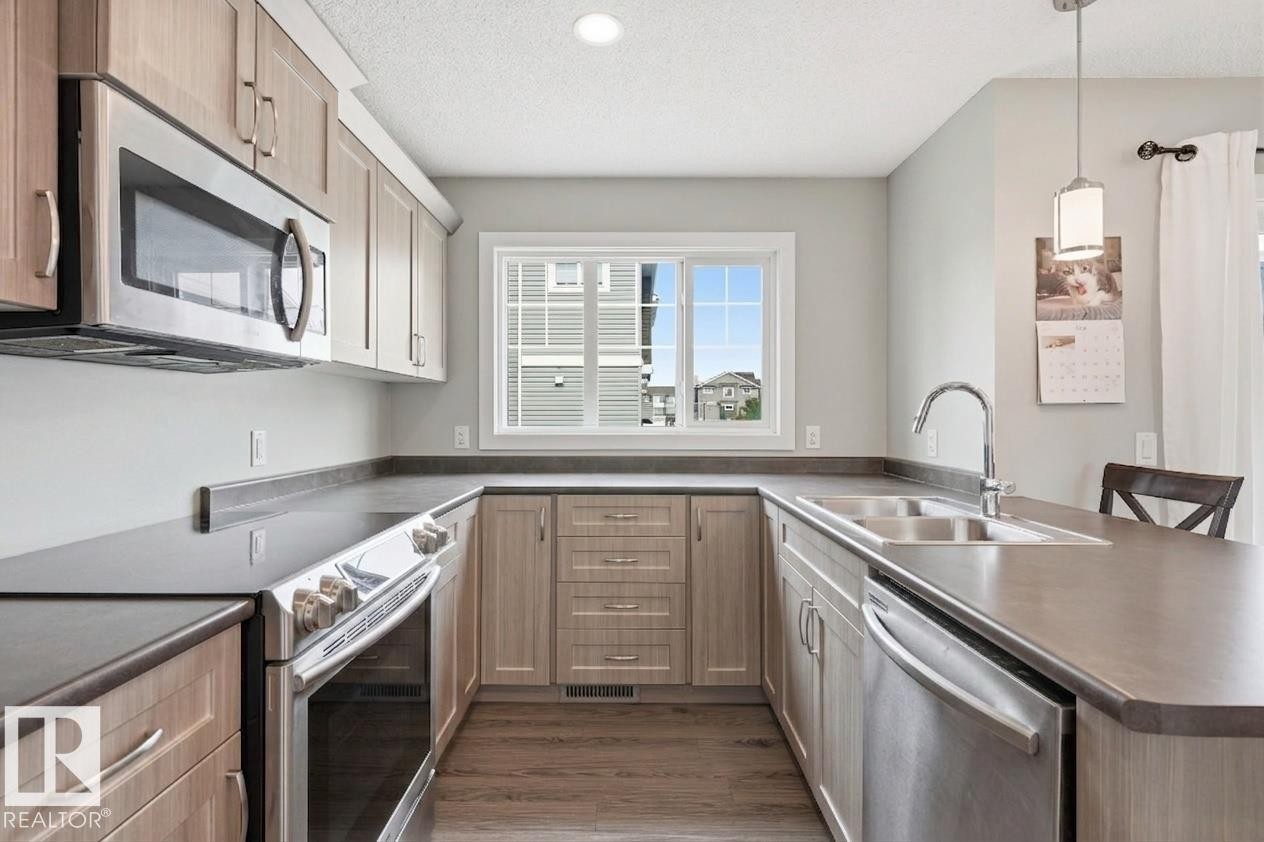 Modern kitchen featuring custom wood-finish cabinetry, stainless steel appliances including a built-in microwave and dishwasher, gray quartz countertops, a chrome single-handle faucet, and wood-finish flooring throughout - #12 1391 Starling Dr Nw, Edmonton, AB