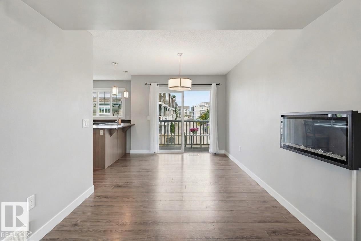 Wood-finish flooring throughout, with a wall-mounted electric fireplace, and a sliding glass door providing access to a balcony with metal railings - #12 1391 Starling Dr Nw, Edmonton, AB