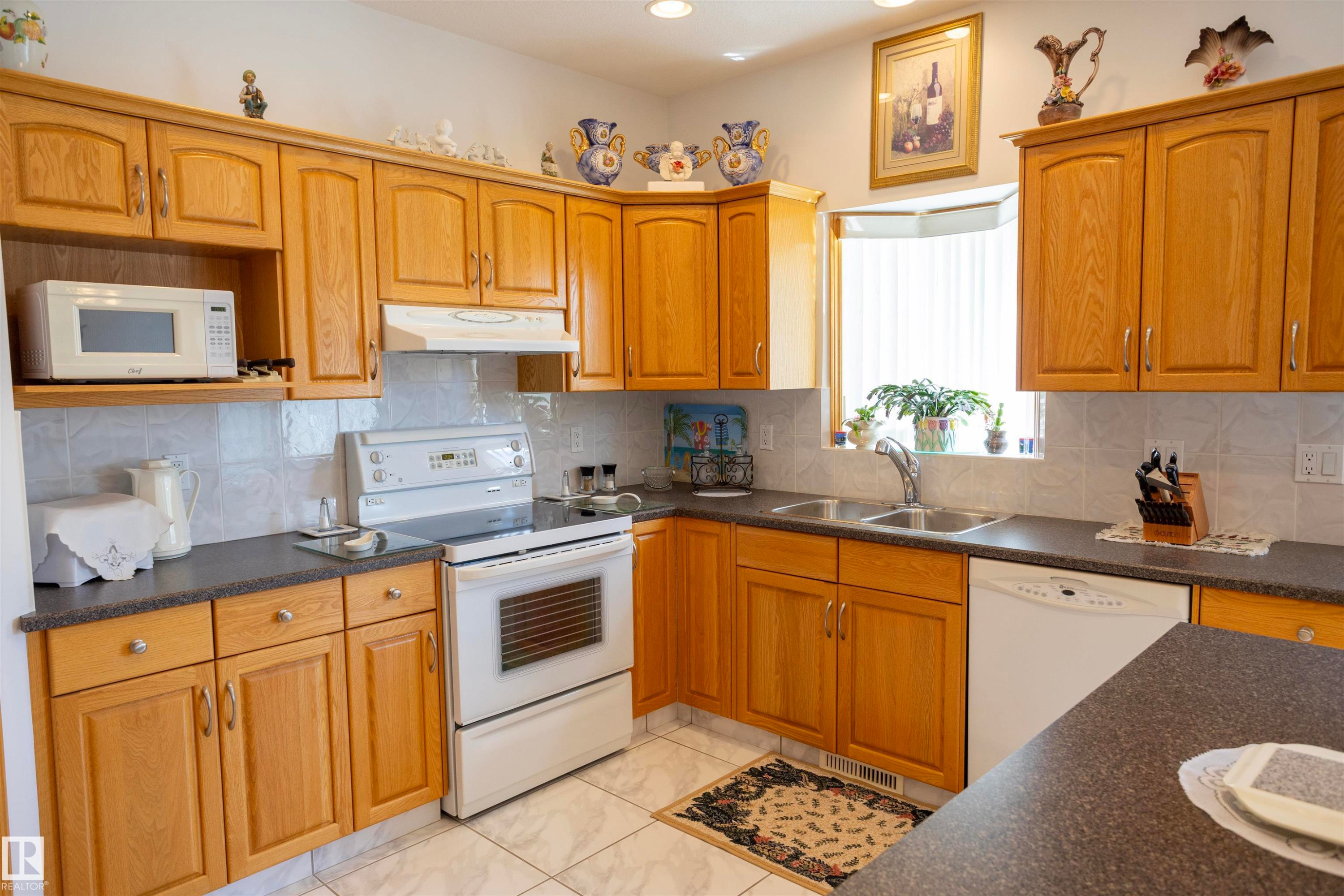 Kitchen featuring abundant wood-finish cabinetry, solid surface countertops, white tile backsplash, and a double basin sink beneath a bay window - 6407 163 Avenue Nw, Edmonton, AB