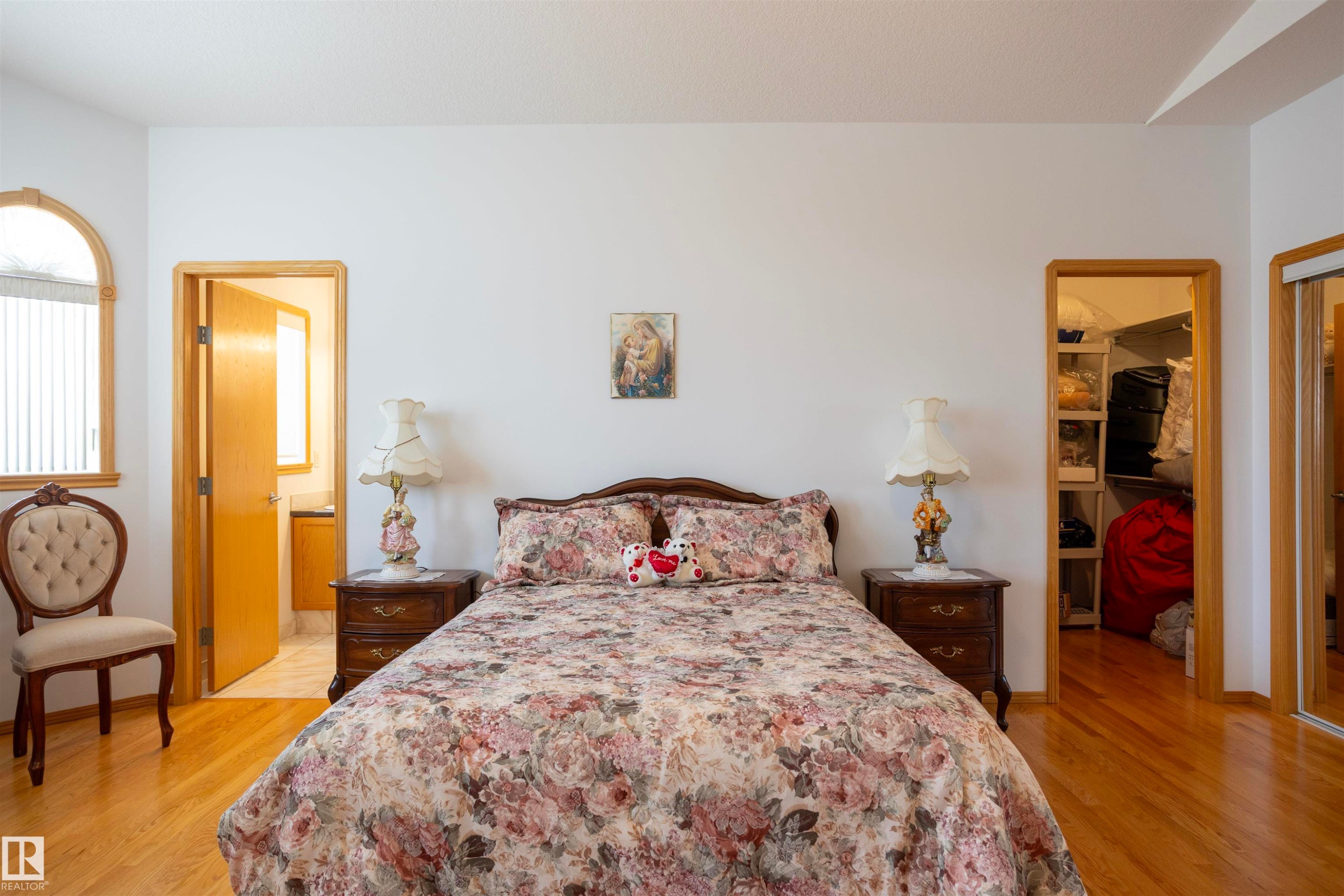 Bedroom featuring light wood-finish flooring, an arched window with blinds, a doorway to a bathroom with a vanity, and a doorway to a closet with built-in shelving - 6407 163 Avenue Nw, Edmonton, AB