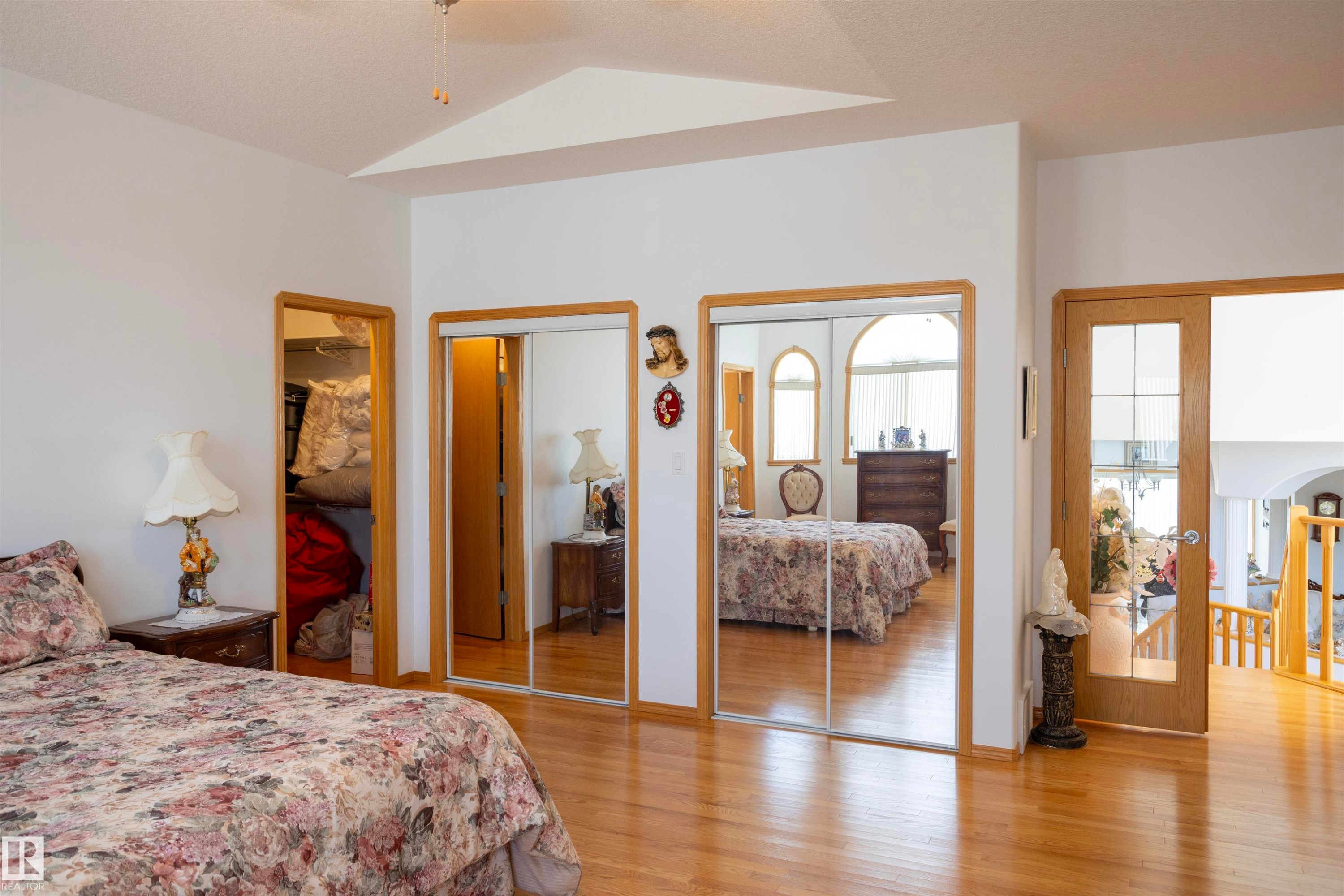 Bedroom featuring wood-finish flooring, two mirrored sliding closet doors, a vaulted ceiling detail, and a wood-frame doorway with glass panels - 6407 163 Avenue Nw, Edmonton, AB