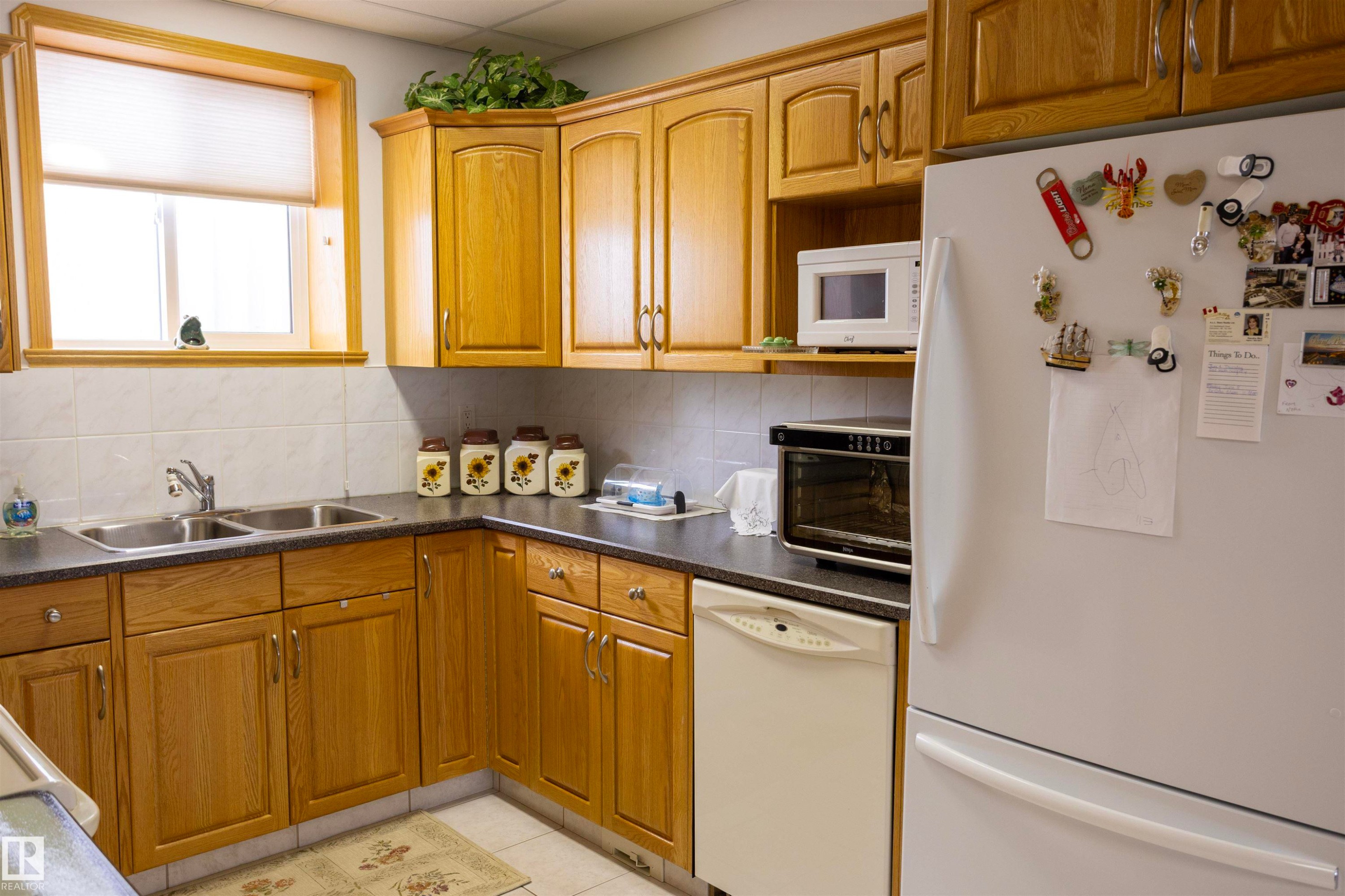 Kitchen featuring light wood cabinetry, a double basin stainless steel sink, dark countertops, white tile backsplash, and a built-in dishwasher - 6407 163 Avenue Nw, Edmonton, AB
