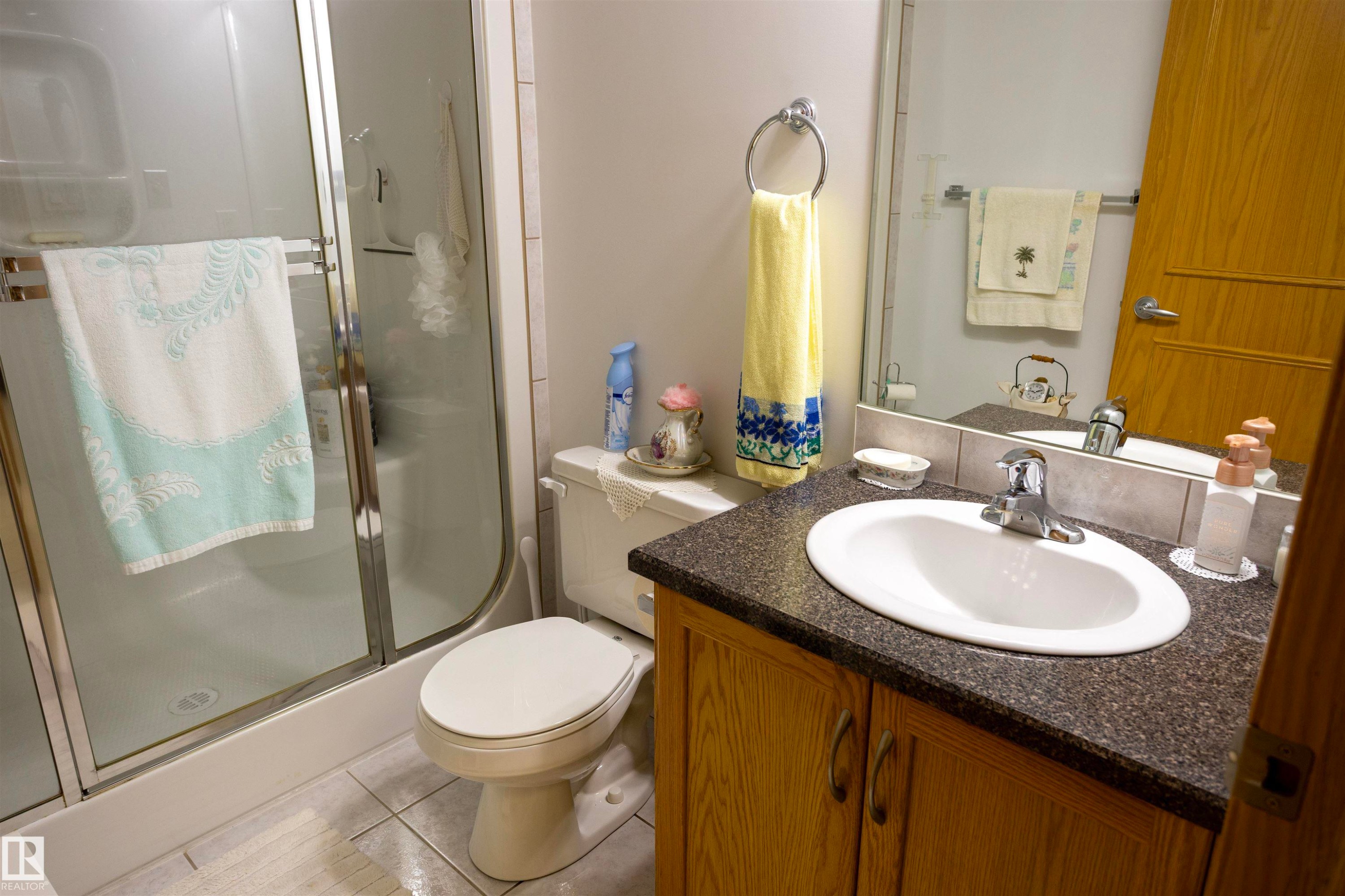 Bathroom featuring a white toilet, enclosed shower with clear glass, wood-finish vanity with a dark countertop, and an oval undermount sink with chrome fixtures - 6407 163 Avenue Nw, Edmonton, AB