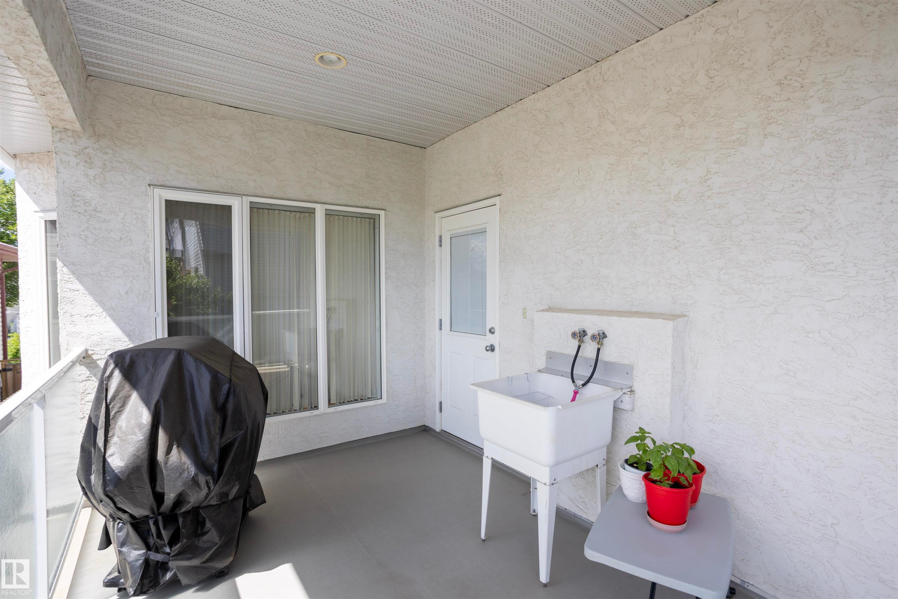 Covered outdoor balcony featuring textured stucco walls, a utility sink with dual faucets, and a white entry door with a window pane - 6407 163 Avenue Nw, Edmonton, AB