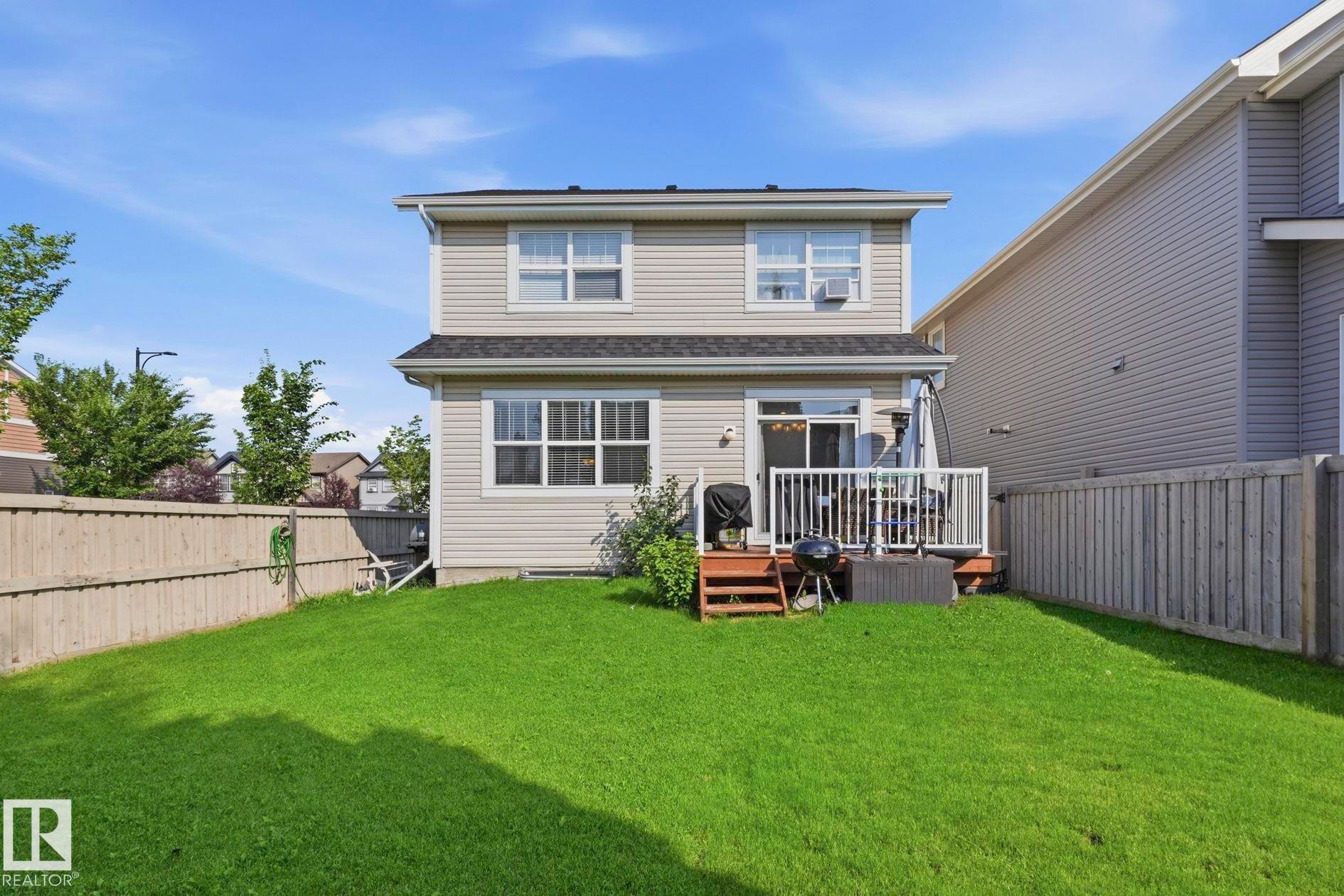 Expansive fenced backyard featuring a manicured lawn, a two-story residence with light-colored siding, and a rear deck with white railings - 3204 Abbott Crescent Sw, Edmonton, AB - Outdoor