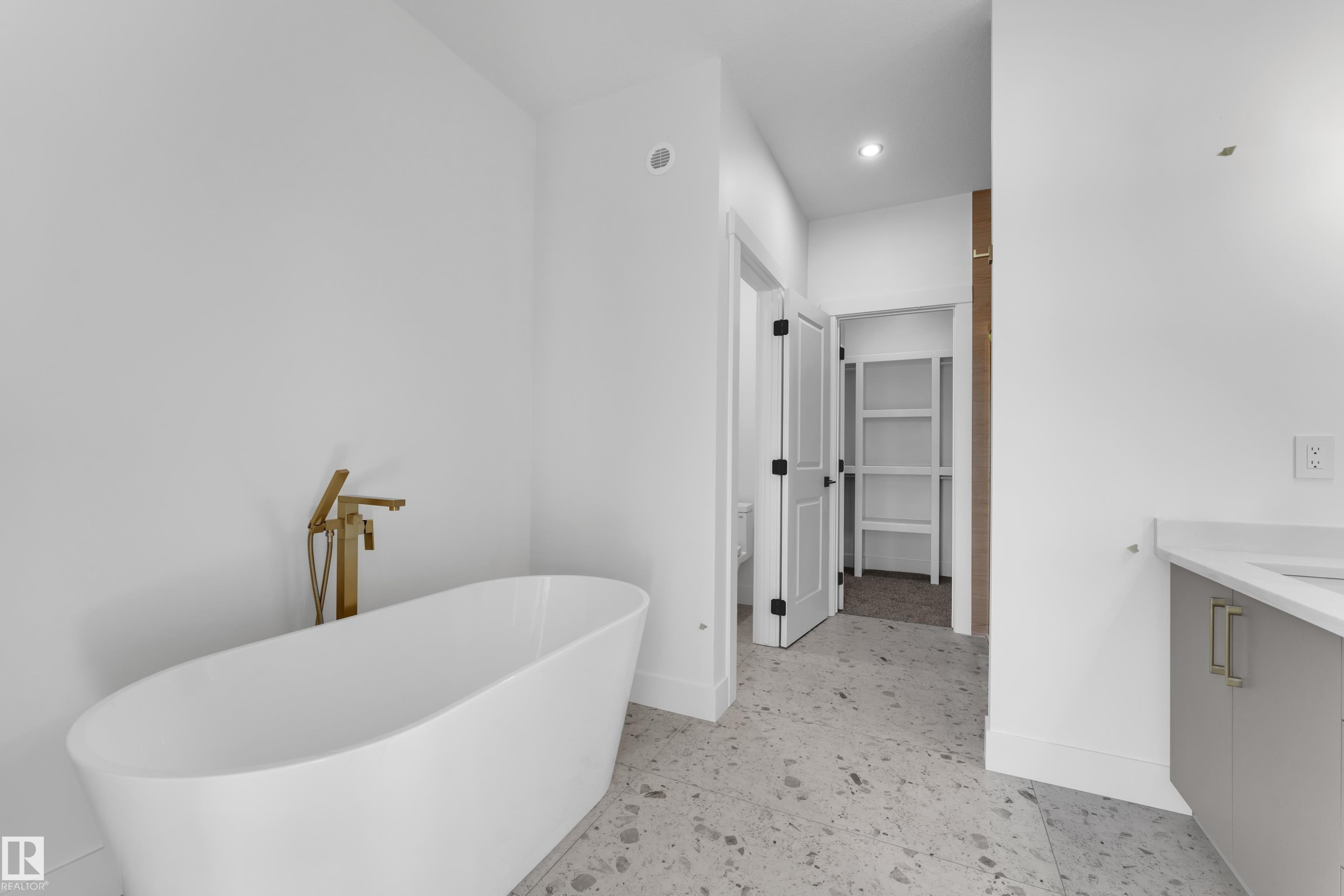 Freestanding soaking tub with a floor-mounted gold-tone faucet, stone-finish flooring, and white walls - 226 Linden Loop, Leduc, AB