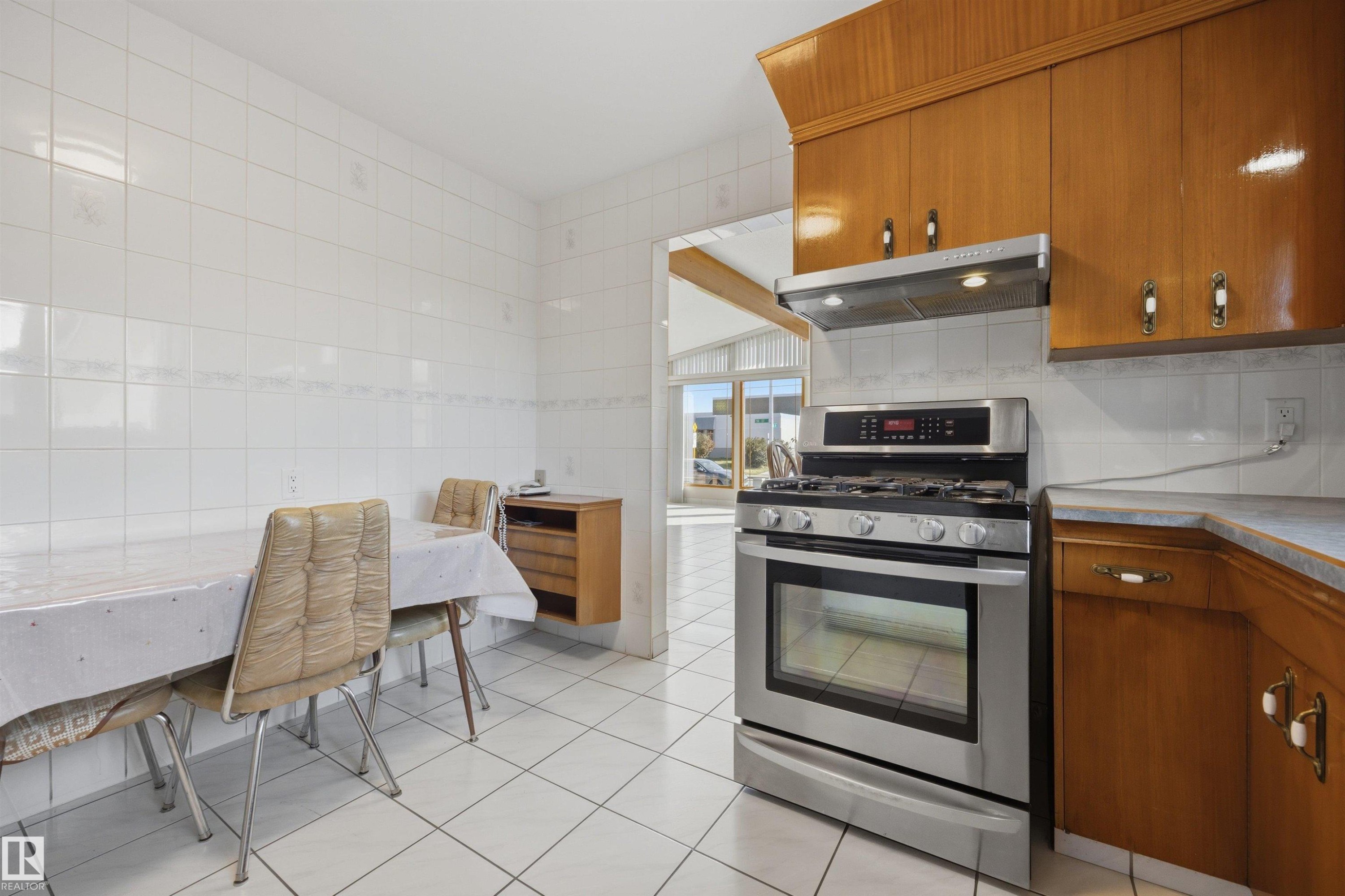 White tiled walls and flooring, wood-finish cabinetry, stainless steel range and oven, stainless steel range hood, and light-toned countertop - 14008 94 Street Nw, Edmonton, AB