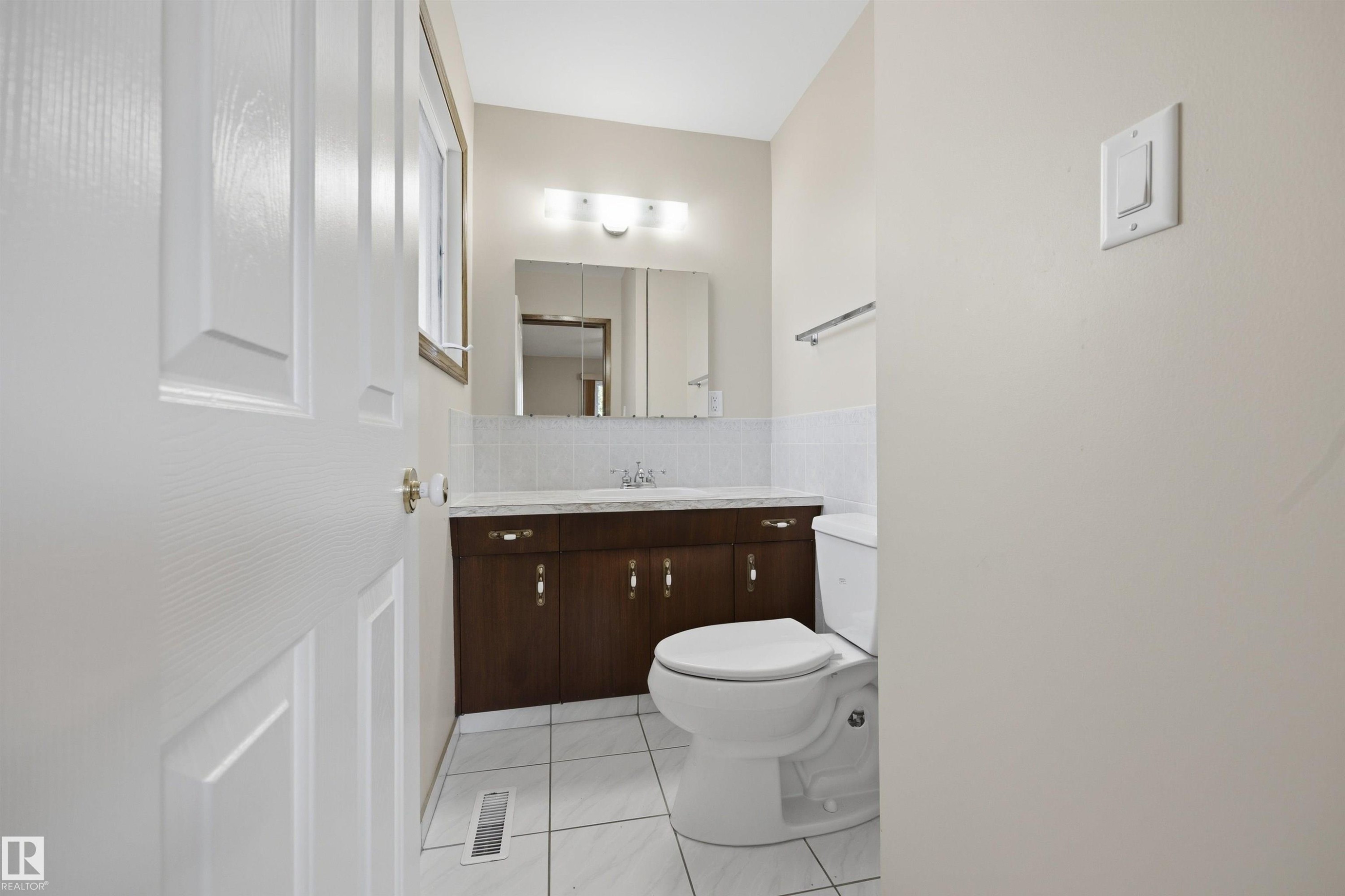 White paneled door, wood-finish vanity with countertop, white tile flooring, wall-mounted mirror, and vanity lighting - 14008 94 Street Nw, Edmonton, AB