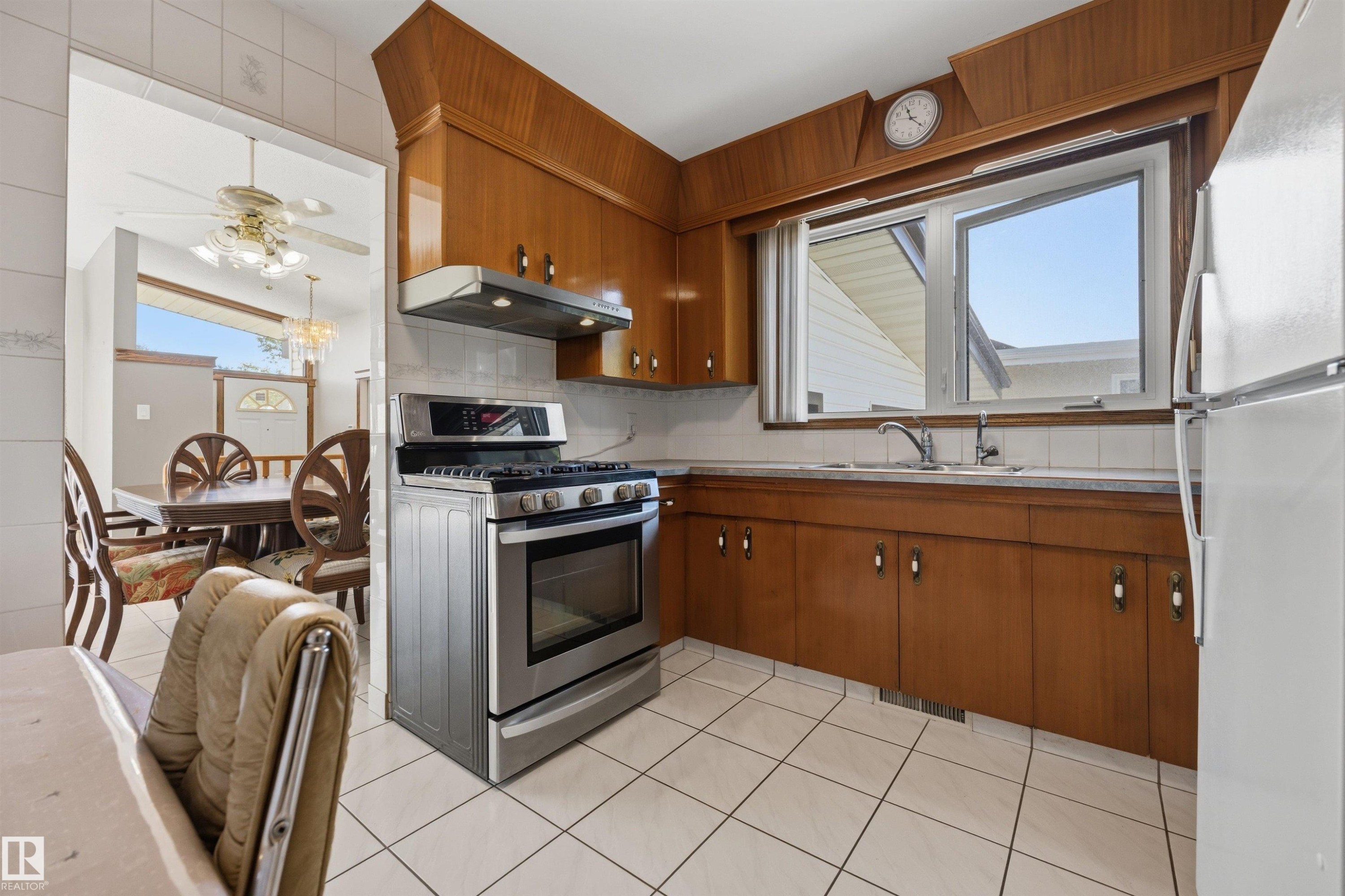 Kitchen featuring wood cabinetry, a stainless steel range with overhead hood, a window over the sink, and white square tile flooring - 14008 94 Street Nw, Edmonton, AB