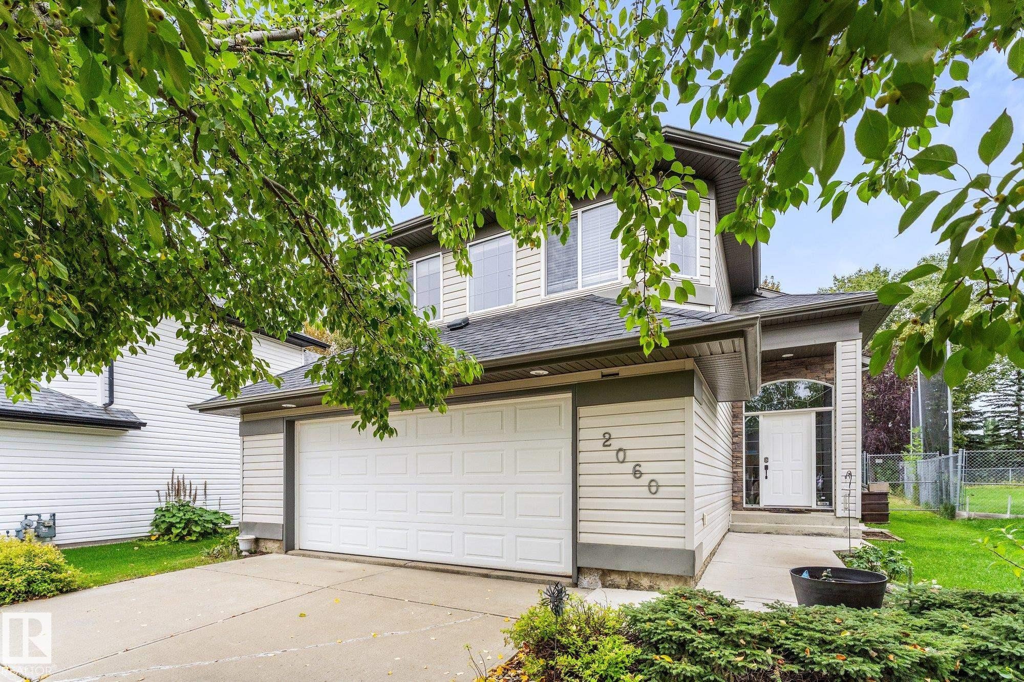 Two-story residence featuring light-toned siding, dark roof shingles, an attached garage, a paved driveway, and a covered entry - 2060 Brennan Crescent Nw, Edmonton, AB