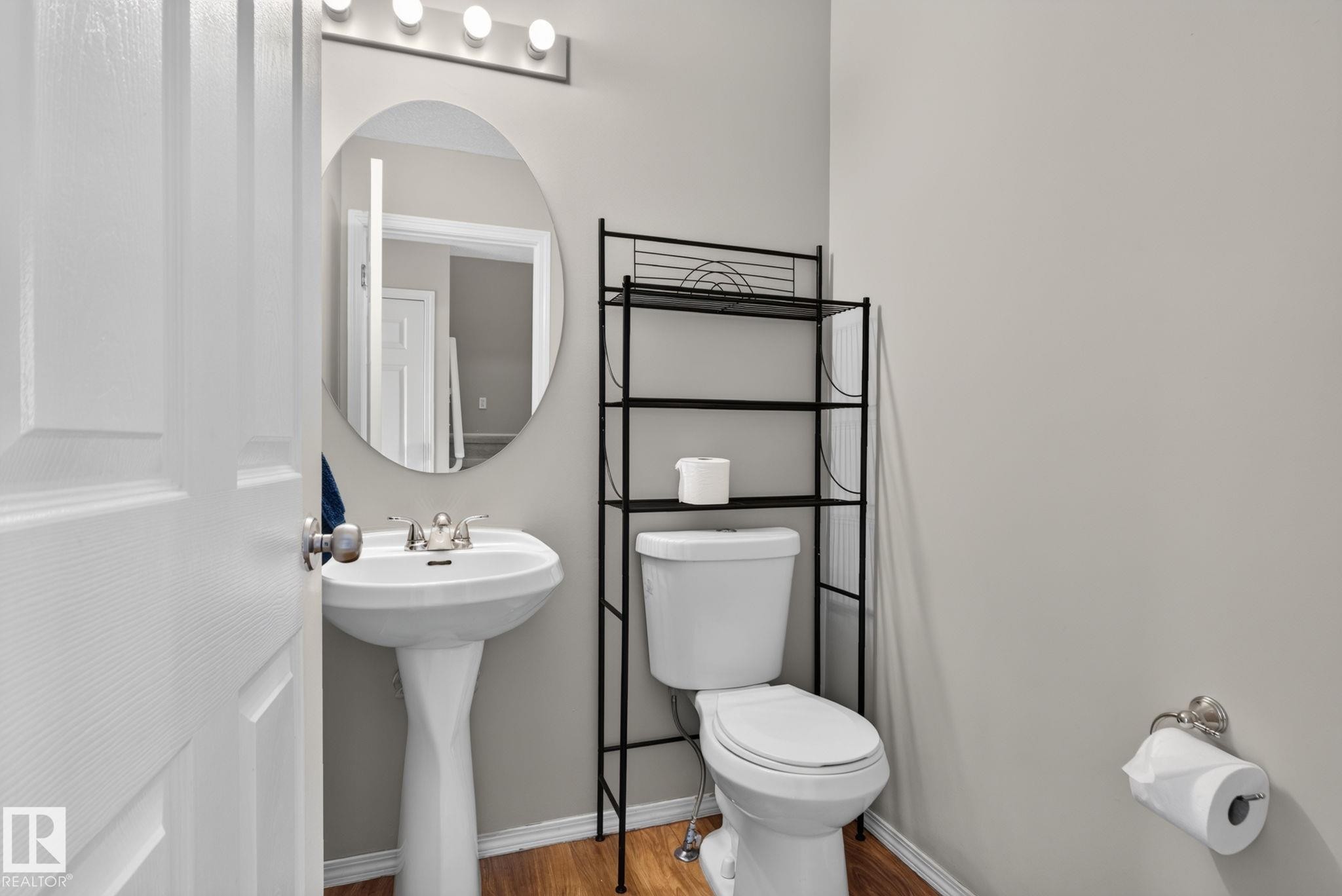 Pedestal sink with chrome faucet, oval wall mirror, modern light fixture, toilet, and wood-finish flooring - 8923 173 Avenue Nw, Edmonton, AB