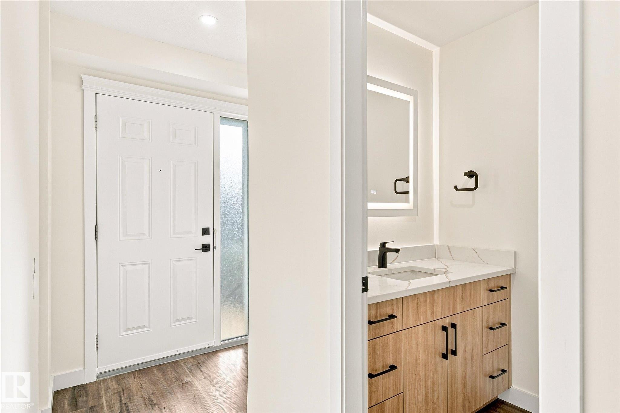 Wood-finish flooring, white paneled entry door with frosted sidelight, wood-tone vanity with a white countertop, and an illuminated mirror - 17325 85 Avenue Nw, Edmonton, AB