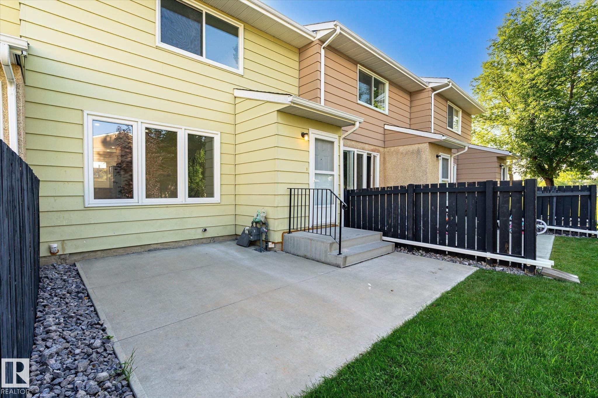 Concrete patio with a black privacy fence, green lawn, and light green siding - 17325 85 Avenue Nw, Edmonton, AB