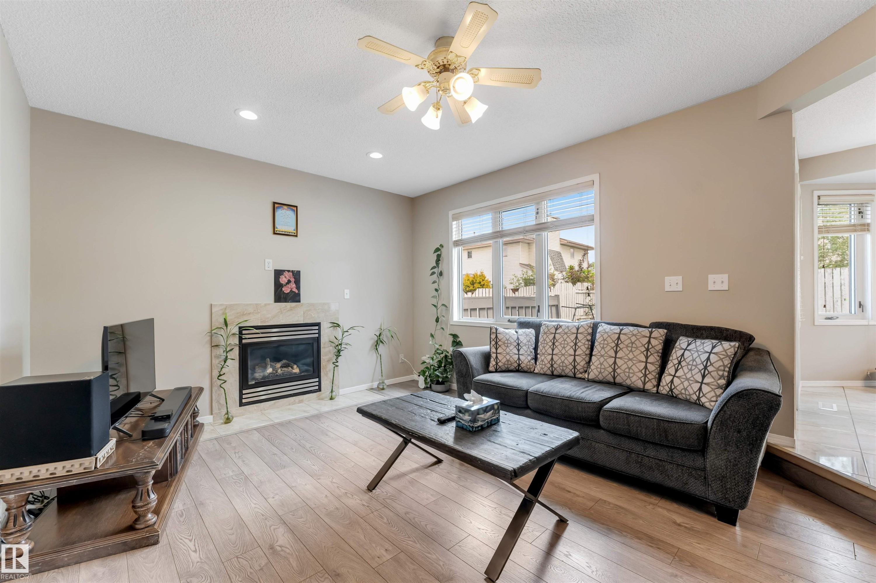 Living area featuring wood-finish flooring, a ceiling fan with integrated lighting, recessed lighting, a fireplace with a stone surround, and windows with blinds - 4465 32A Street Nw, Edmonton, AB - Indoor Photo Showing Living Room With Fireplace