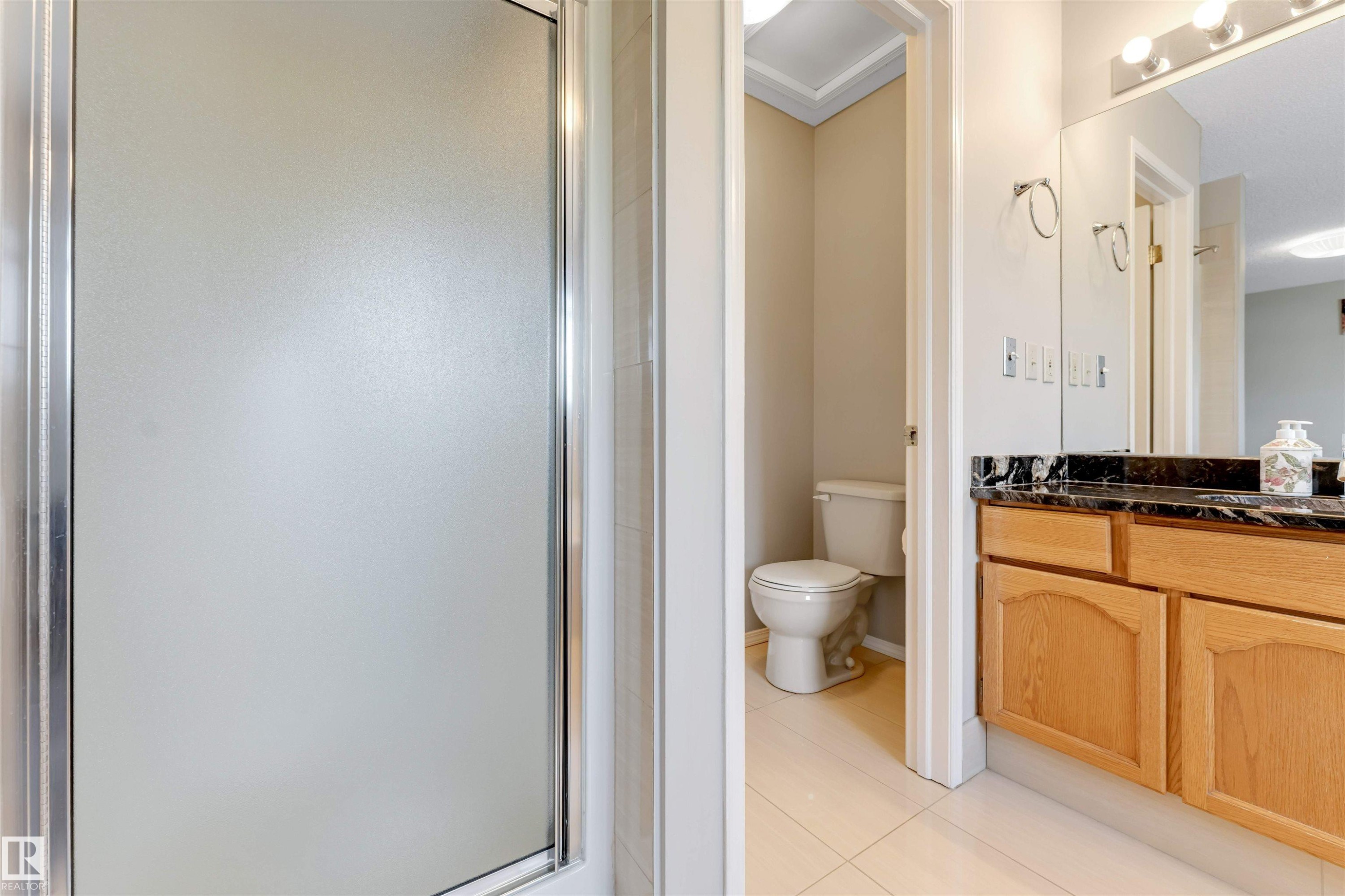 Shower enclosure with frosted glass and chrome framing, complemented by a wood-finish vanity featuring a dark stone countertop, a large framed mirror, and a separate commode area - 4465 32A Street Nw, Edmonton, AB - Indoor Photo Showing Bathroom