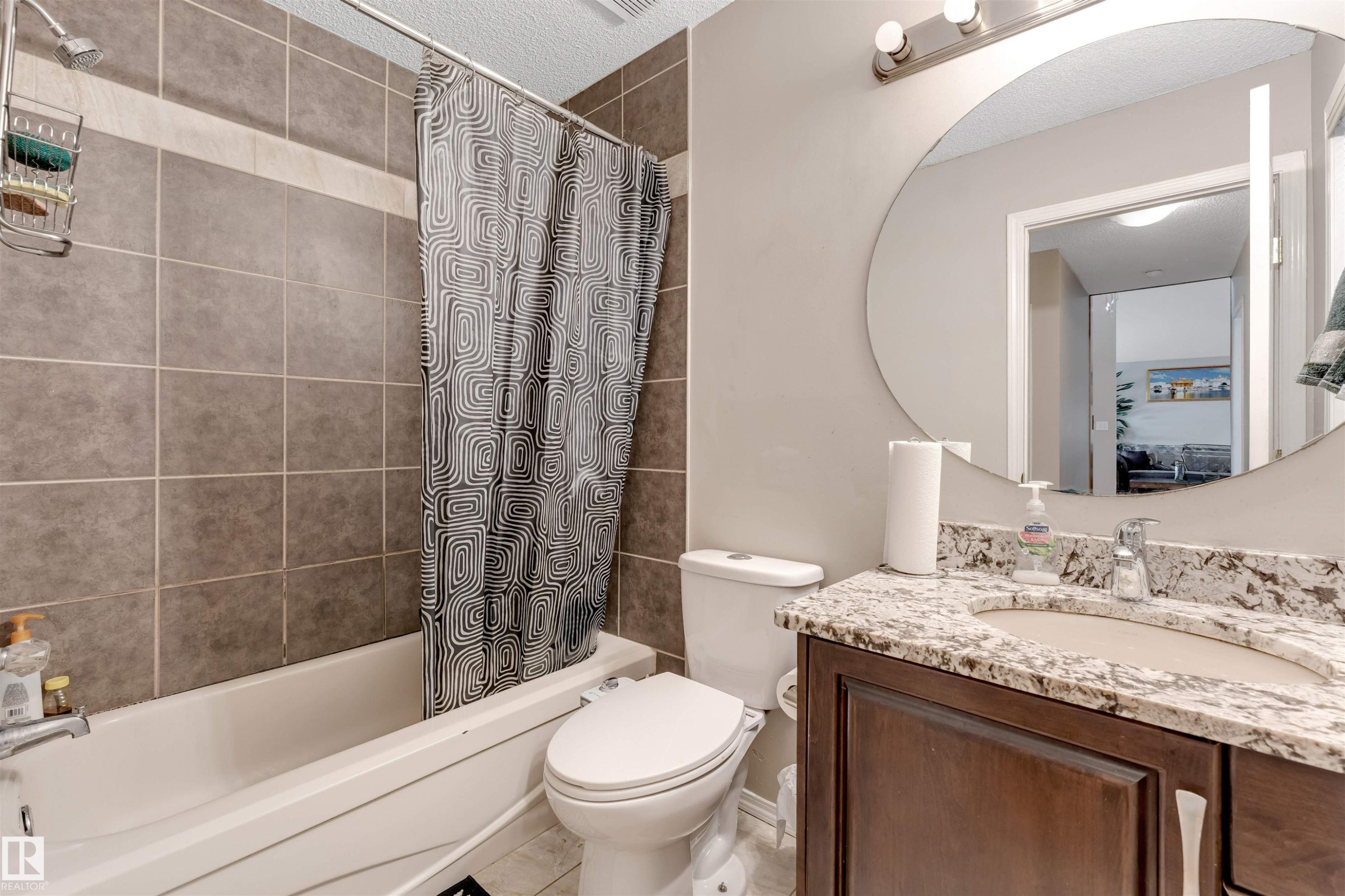 Bathroom featuring a tub/shower combination with tile surround, a dark wood-finish vanity with a stone-look countertop, a circular mirror, and overhead vanity lighting - 4465 32A Street Nw, Edmonton, AB - Indoor Photo Showing Bathroom