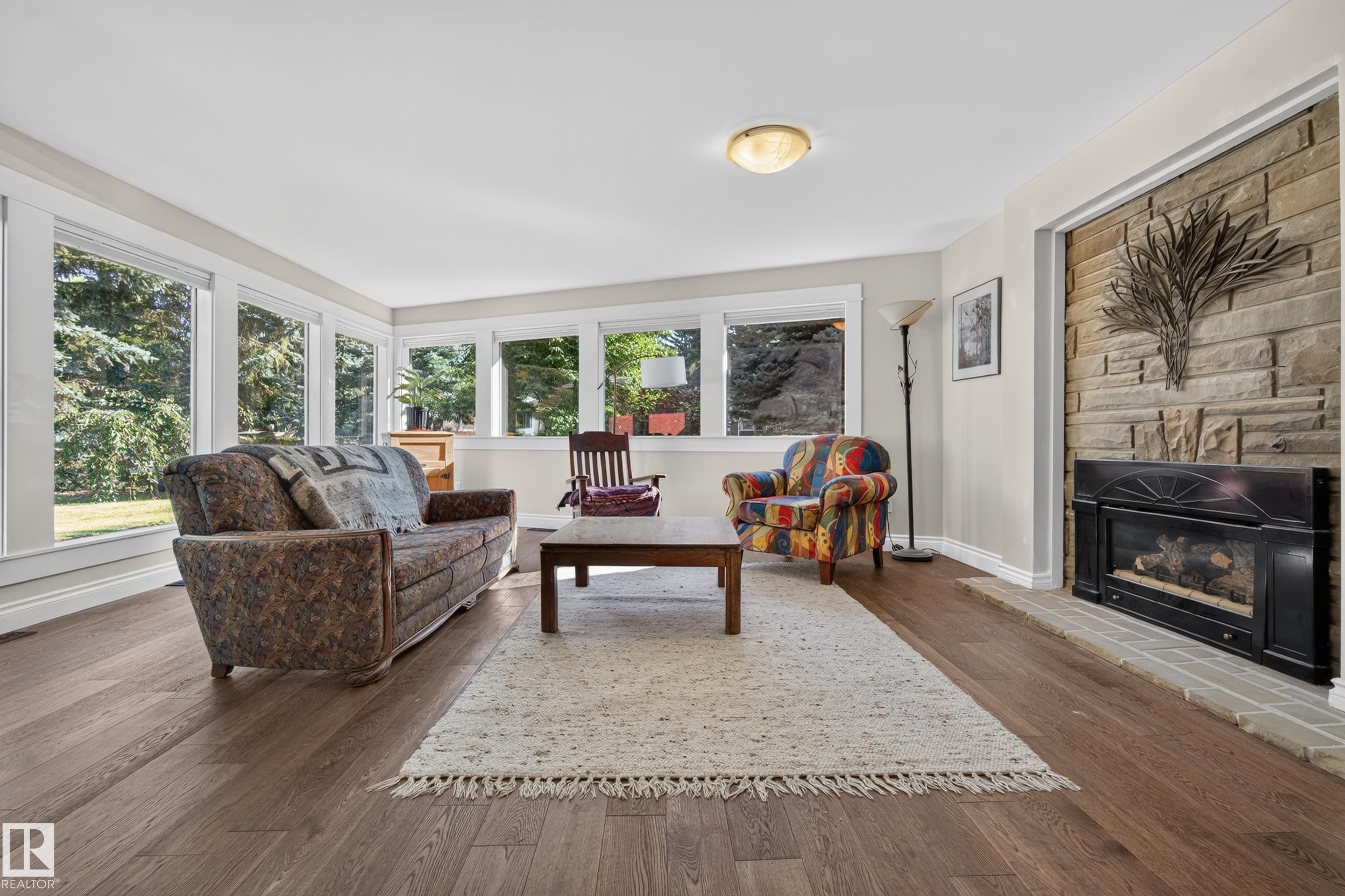 Wood-finish flooring, stone-clad fireplace with a black insert, white baseboards, and multiple large windows - 11603 72 Avenue Nw, Edmonton, AB