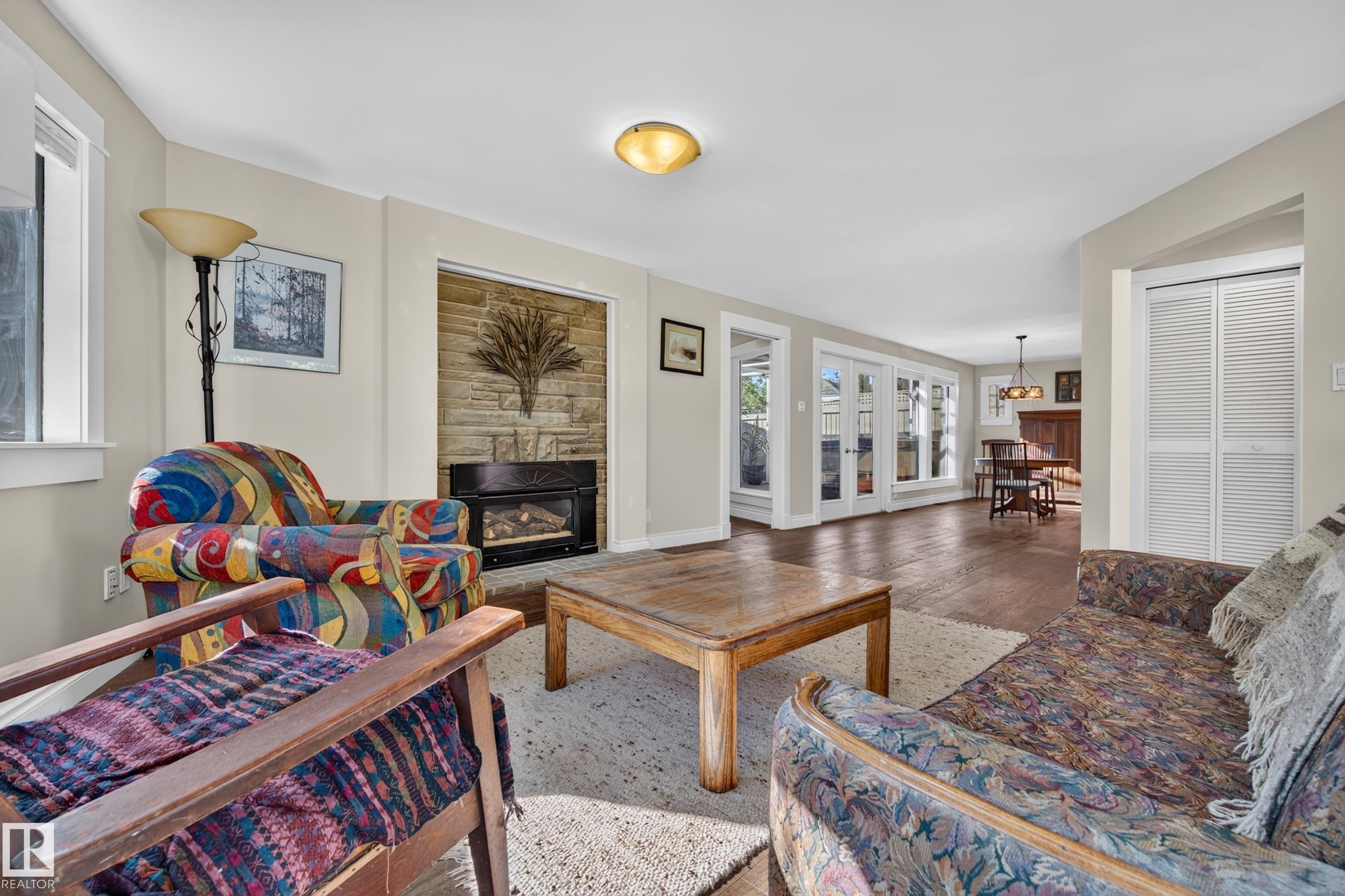 Stone-faced fireplace with an inset gas insert, wood-finish flooring, white louvered bifold doors, and glass-paned French doors - 11603 72 Avenue Nw, Edmonton, AB
