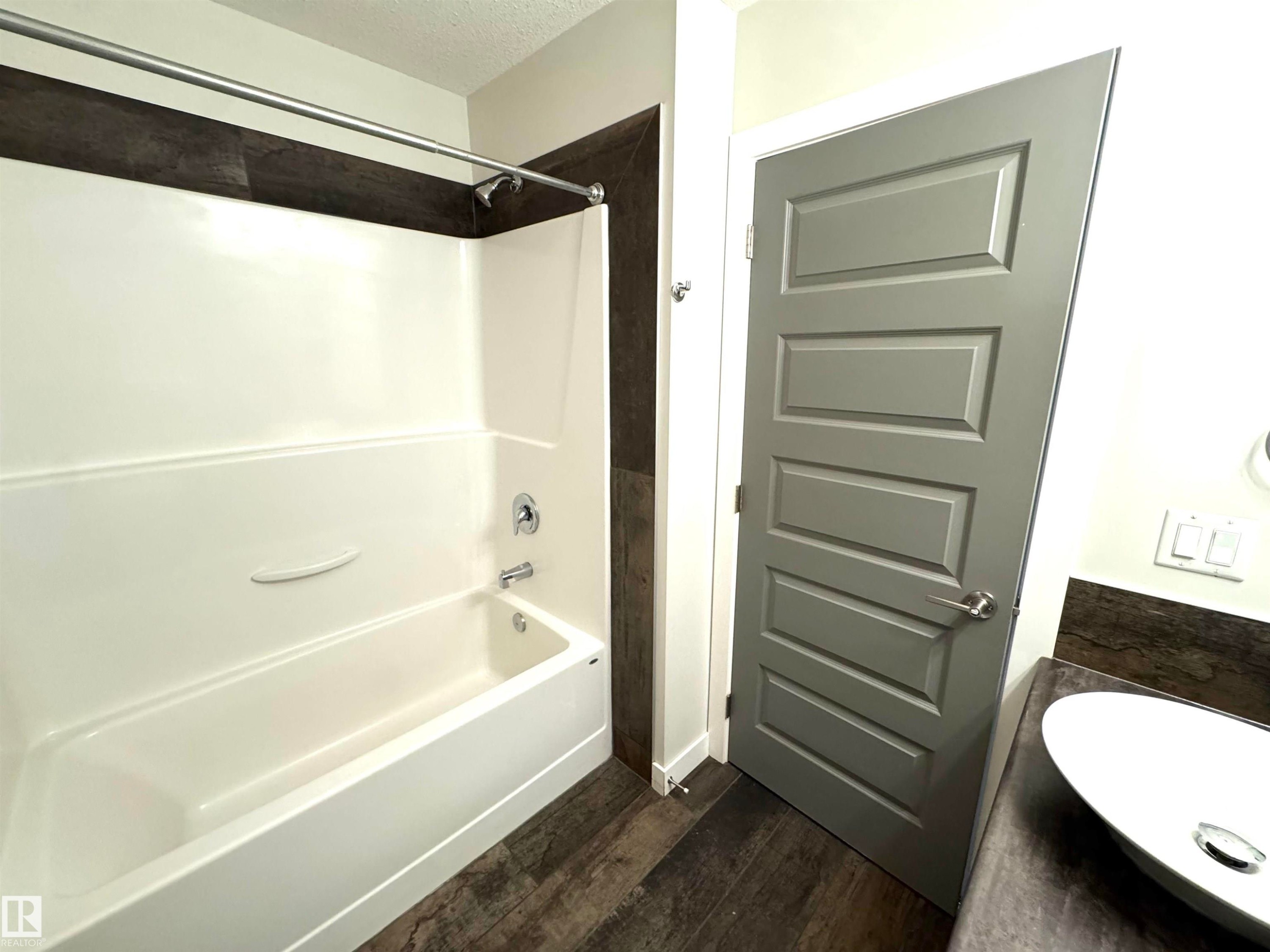 White bathtub with chrome fixtures, dark wood-finish flooring, and a gray paneled door - 12046 89 Street Nw, Edmonton, AB
