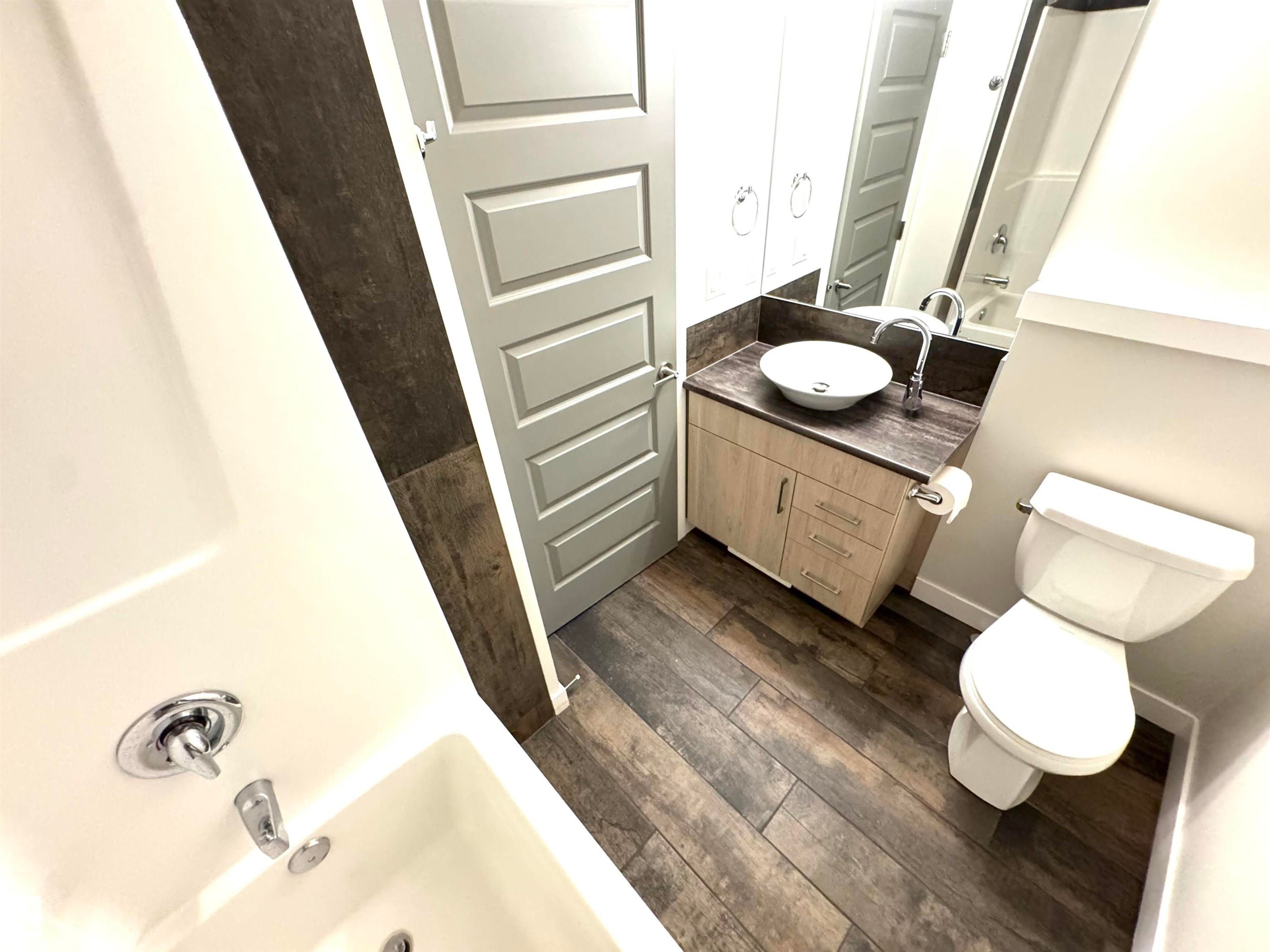 Wood-finish plank flooring, vanity with light wood-finish cabinetry, vessel sink with chrome faucet, full-width mirror, and a white bathtub with chrome fixtures - 12046 89 Street Nw, Edmonton, AB