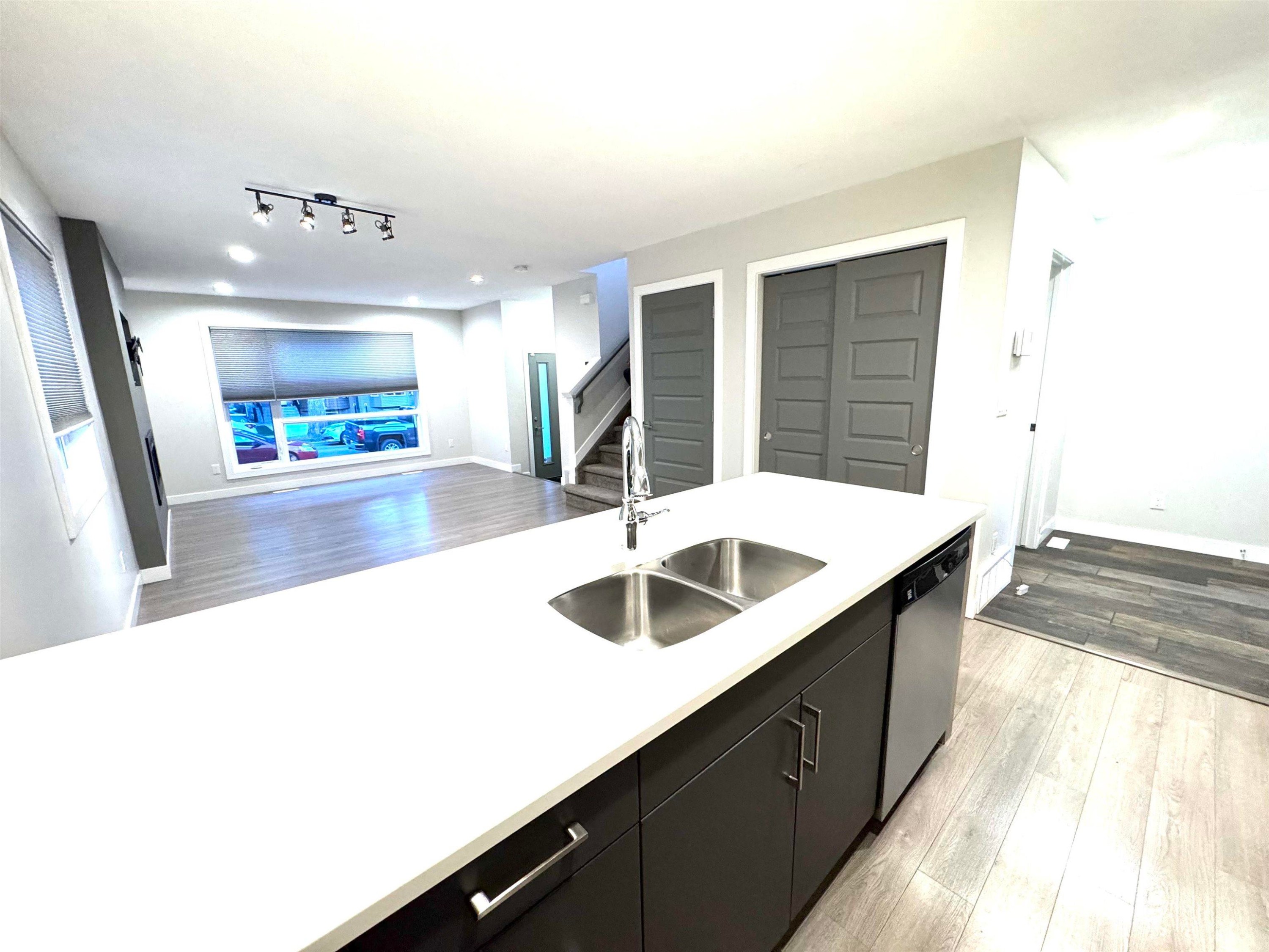 Open-concept living space featuring wood-finish flooring, a large window, a track lighting fixture, and a kitchen island with a double stainless steel sink - 12046 89 Street Nw, Edmonton, AB