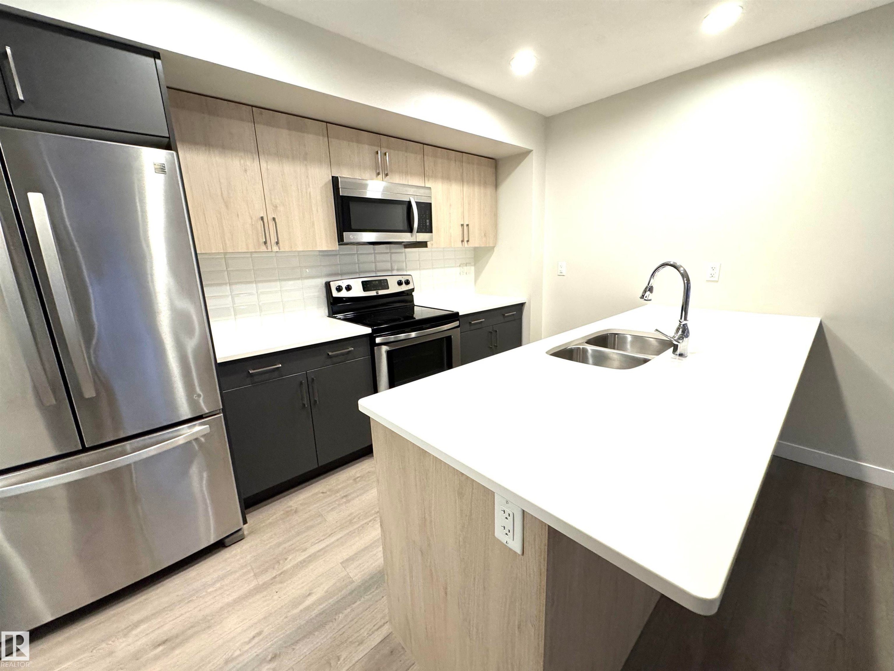 Modern kitchen featuring stainless steel appliances, dual-tone cabinetry, wood-finish flooring, a white countertop island with an integrated double sink, and a tiled backsplash - 12046 89 Street Nw, Edmonton, AB