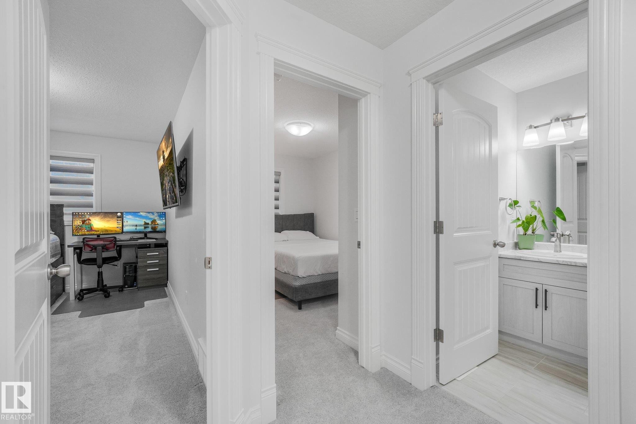 Gray carpeted hallway with white walls and trim, leading to a bathroom with a vanity and a separate room with a bed - 12843 67 Street Nw, Edmonton, AB