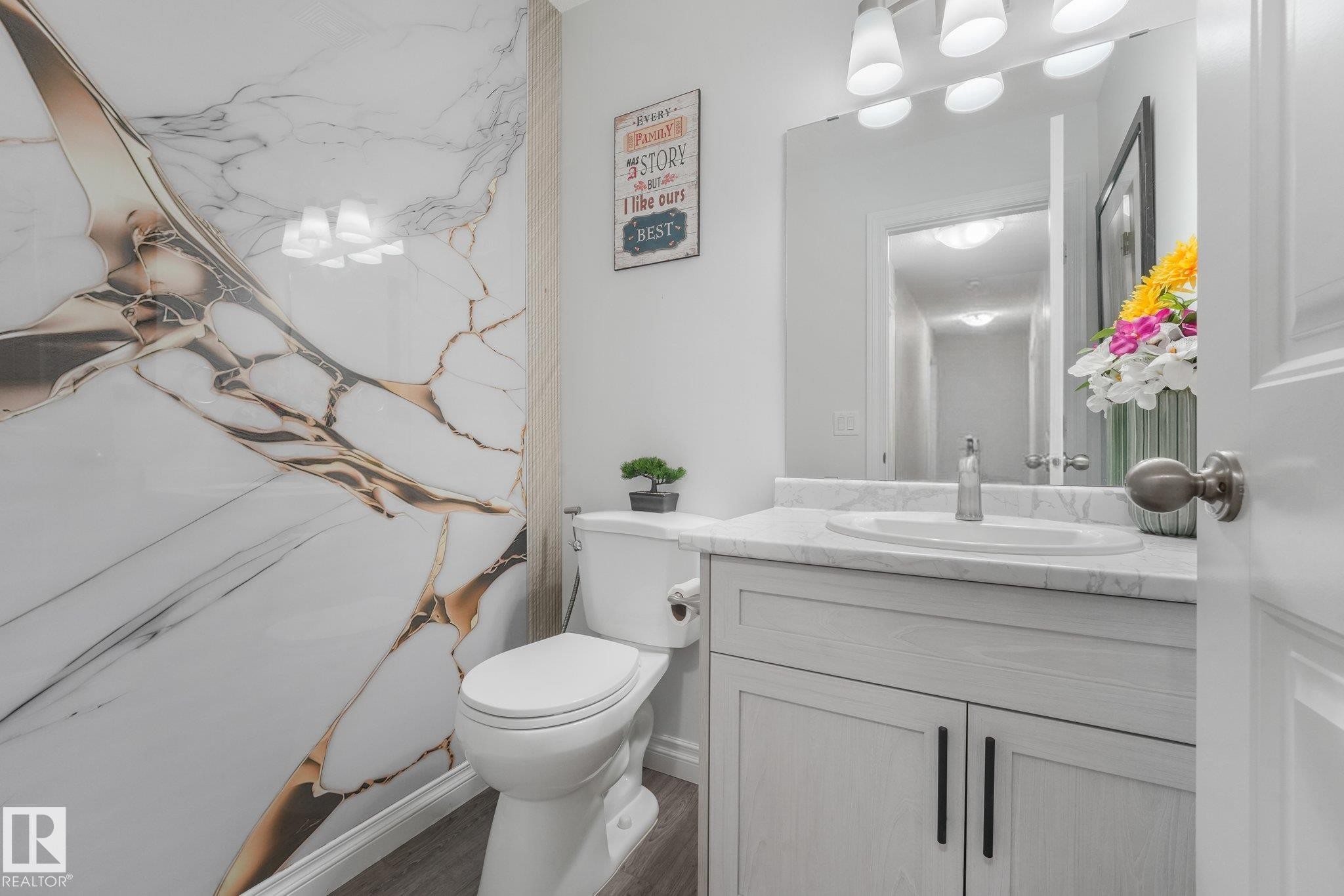 Vanity with a white countertop and integrated sink, under-sink cabinetry with black hardware, wall-mounted mirror, and a wall panel with gold-toned veining - 12843 67 Street Nw, Edmonton, AB