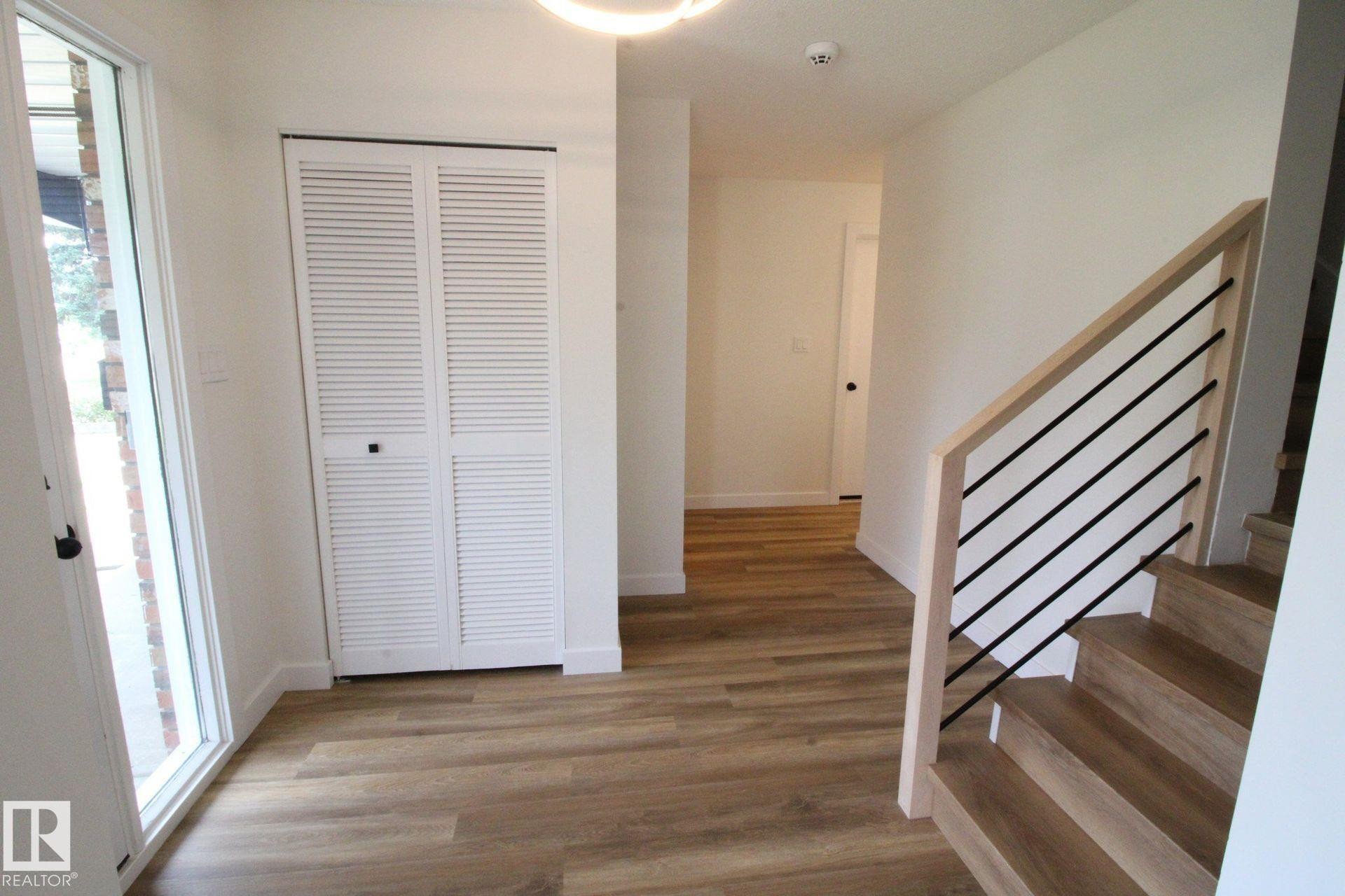 Wood-finish flooring throughout, white louvered bifold closet doors, and a staircase with light wood banister and black metal spindles - 4224 110 Street Nw, Edmonton, AB