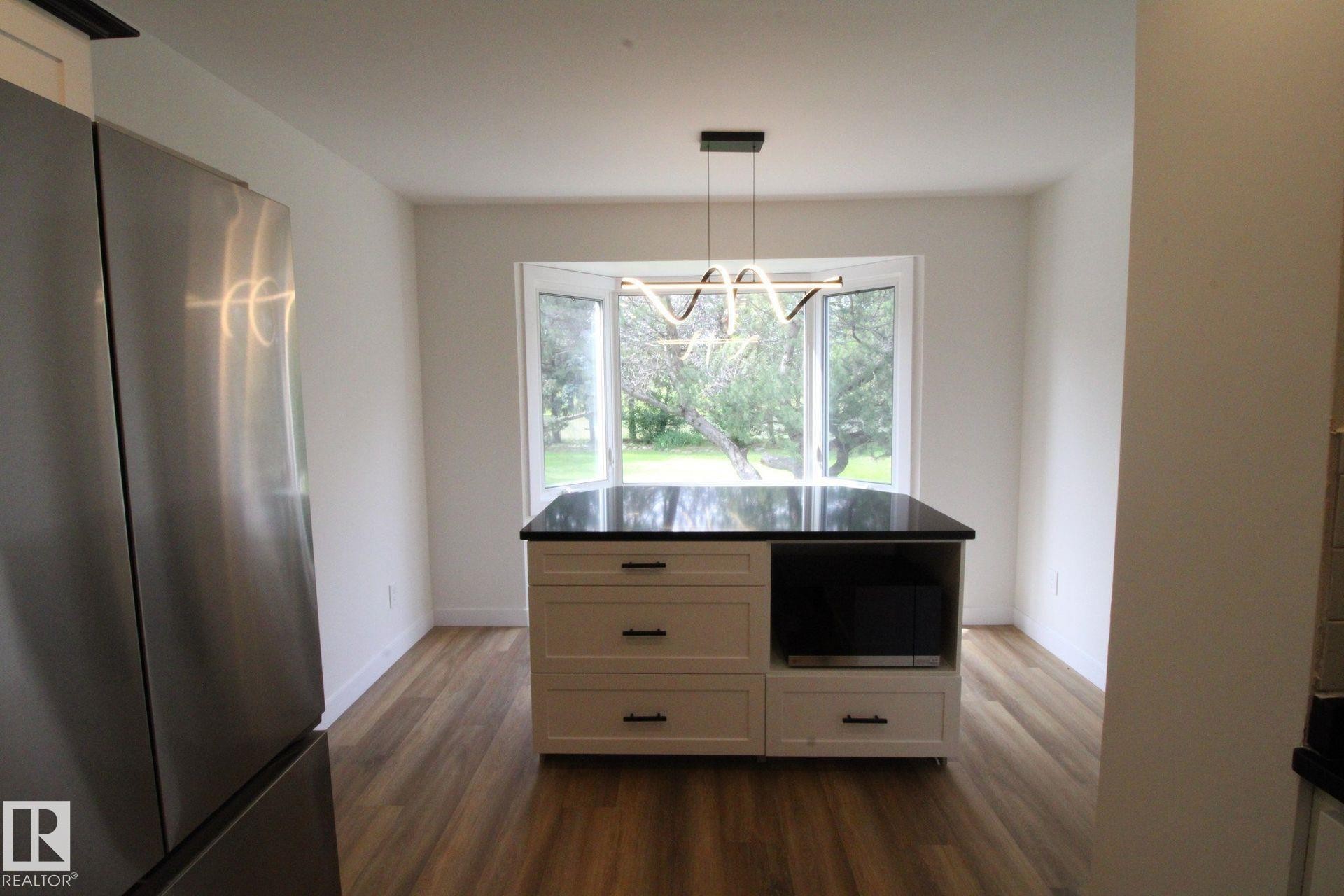 Kitchen featuring a central island with a dark countertop and white cabinetry, wood-finish flooring, a stainless steel refrigerator, and a bay window - 4224 110 Street Nw, Edmonton, AB