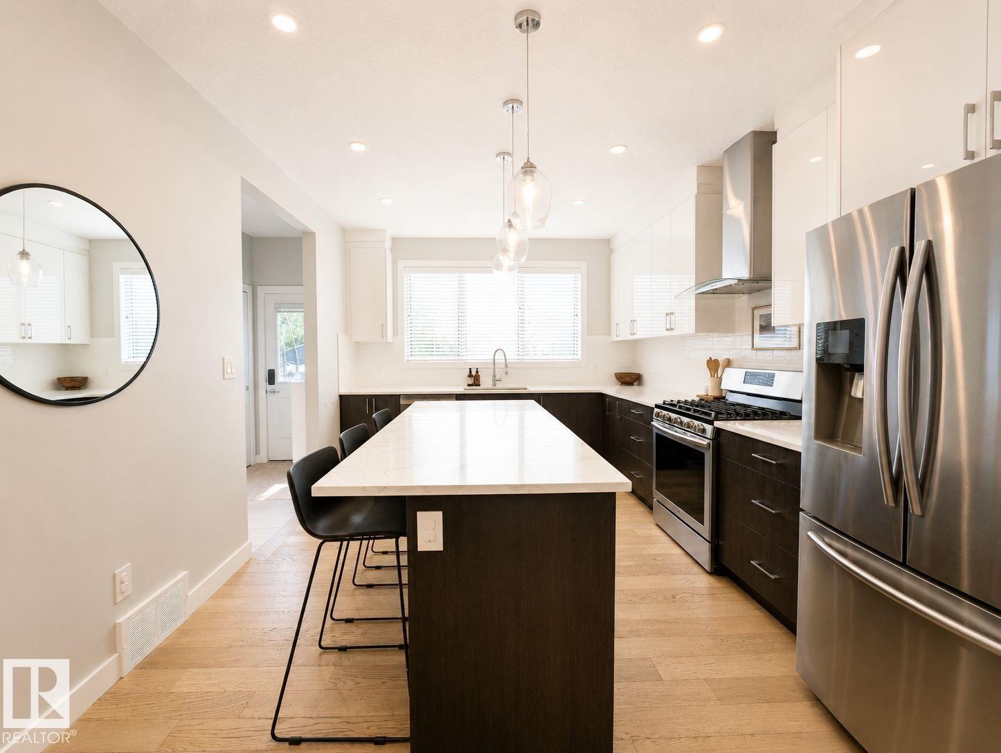 Kitchen featuring a center island with seating, stainless steel appliances, wood-finish flooring, white upper cabinetry, and dark lower cabinetry - 10516 76 Street Nw, Edmonton, AB