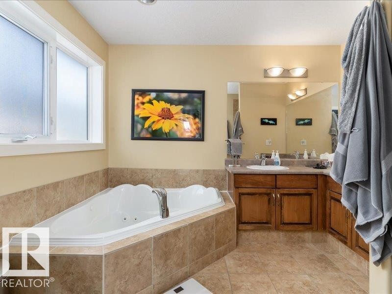 Corner jetted tub with tile surround, adjacent window, and a vanity with a single sink, wood-finish cabinetry, and a large mirror - 496 Ravine Court, Devon, AB