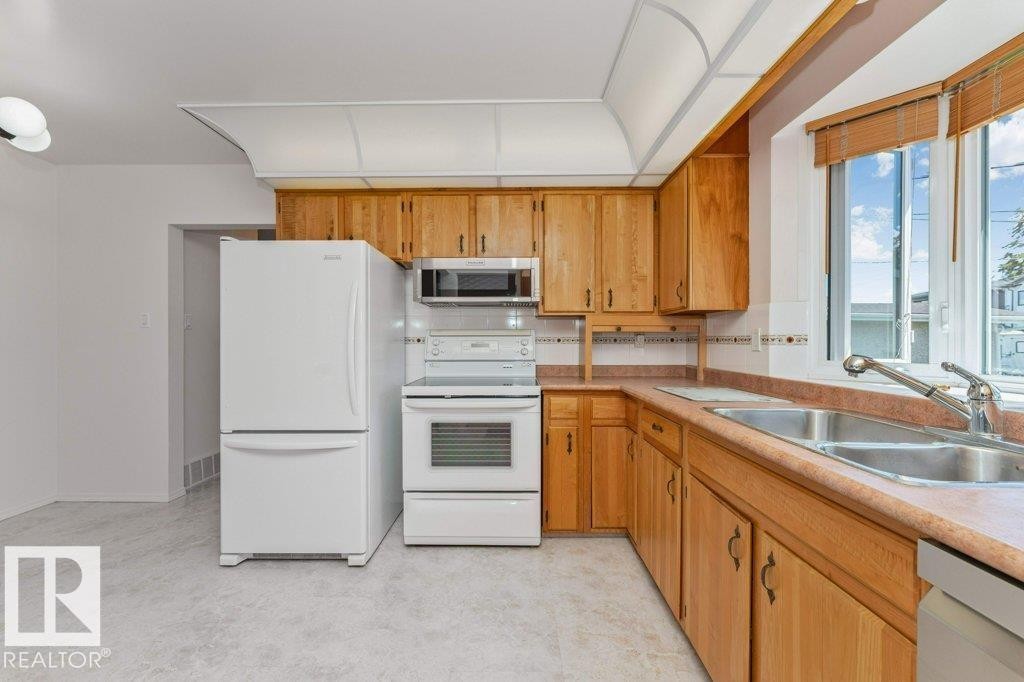 Functional kitchen featuring wood cabinetry, white appliances, dual basin sink, and ample natural light from a large window - 11415 56 Avenue Nw, Edmonton, AB