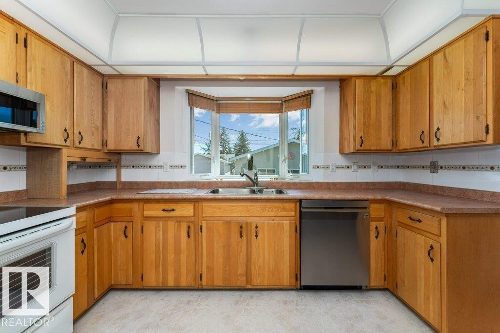 Kitchen featuring abundant wood-finish cabinetry, a bay window with a stainless steel sink, tile backsplash, and a built-in dishwasher - 11415 56 Avenue Nw, Edmonton, AB