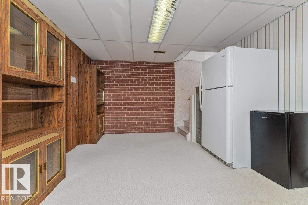 Lower-level room featuring wood-finish cabinetry with glass inserts, exposed brick accent wall, drop ceiling, and carpet flooring - 11415 56 Avenue Nw, Edmonton, AB