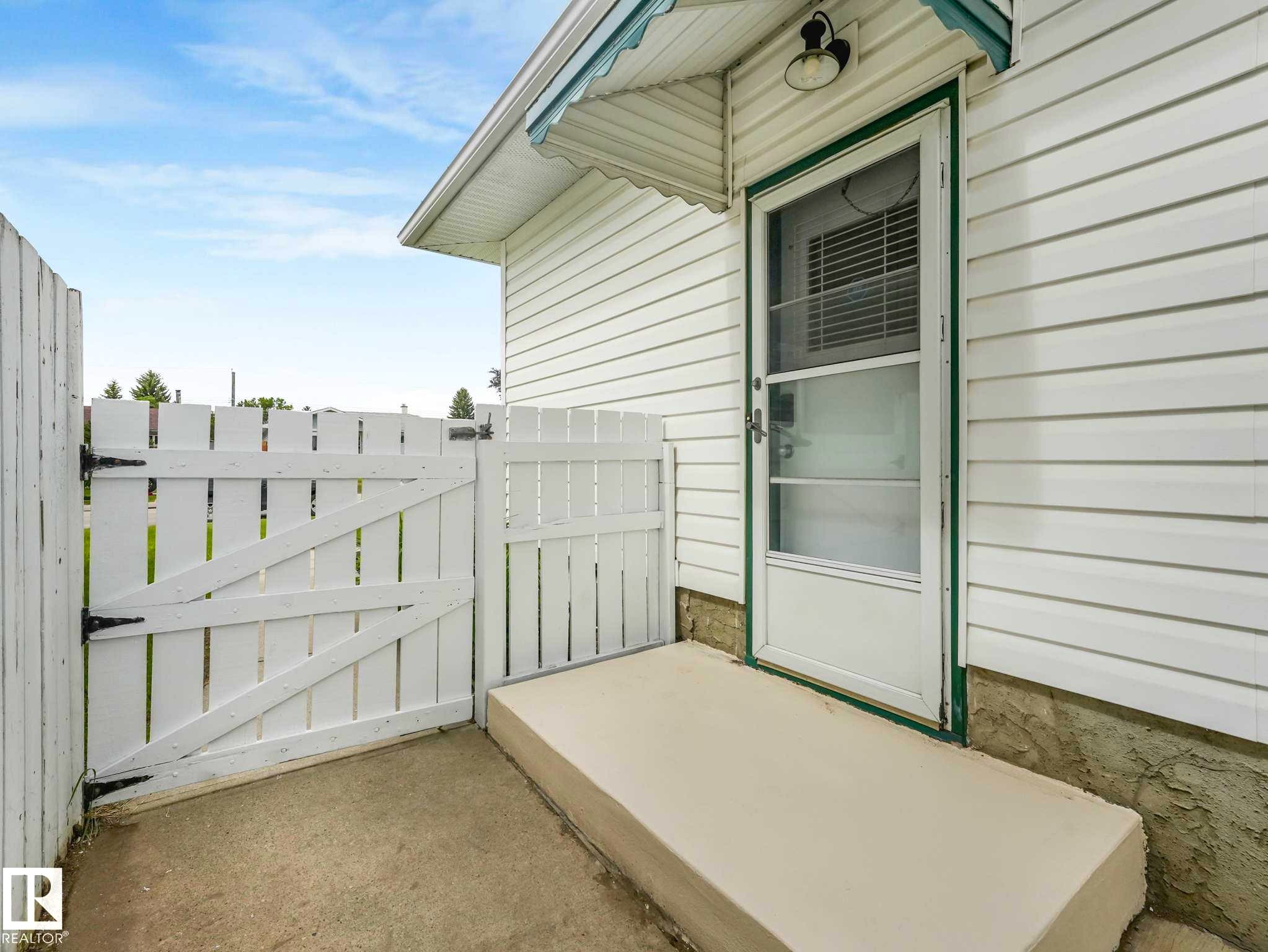 White picket fence with gate, concrete patio, white vinyl siding, exterior lighting fixture, and a storm door - 15916 88B Avenue Nw, Edmonton, AB
