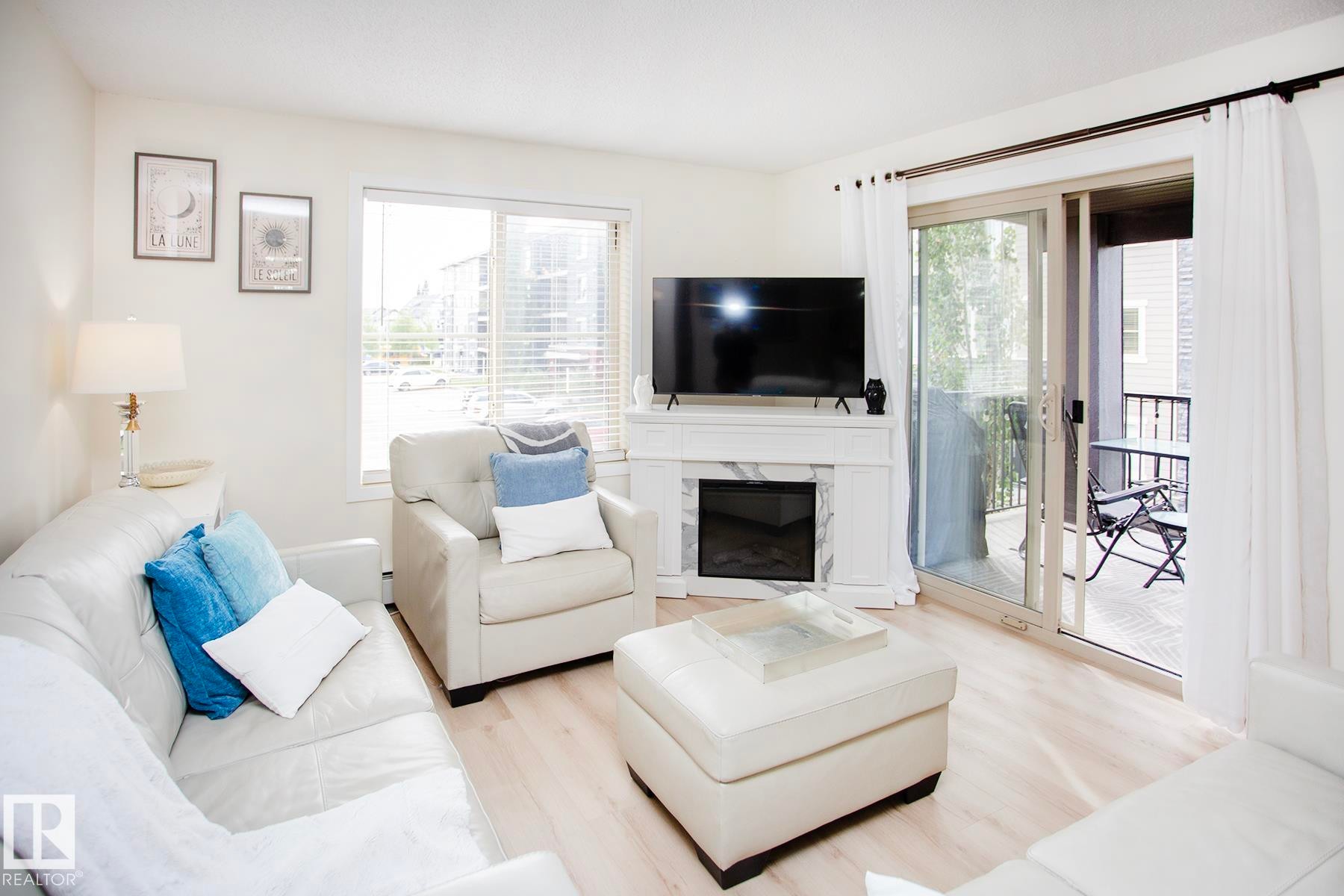 Bright living area featuring wood-finish flooring, a white mantel with a faux fireplace insert, and a sliding glass door opening to an outdoor balcony - #201 1070 Mcconachie Bv Nw, Edmonton, AB