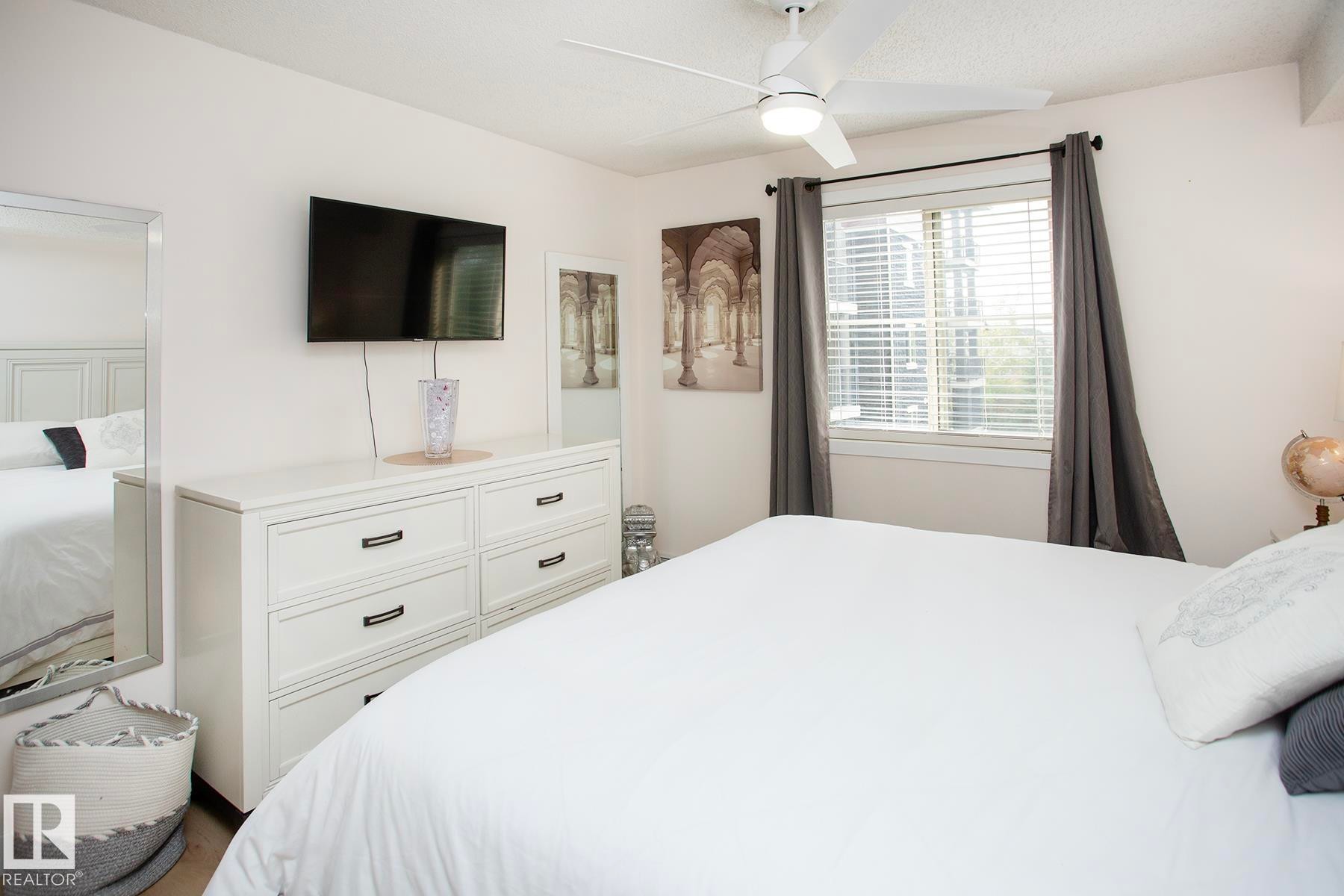 Recessed window with blinds, ceiling fan with integrated lighting, wall-mounted television, and wood-finish flooring - #201 1070 Mcconachie Bv Nw, Edmonton, AB