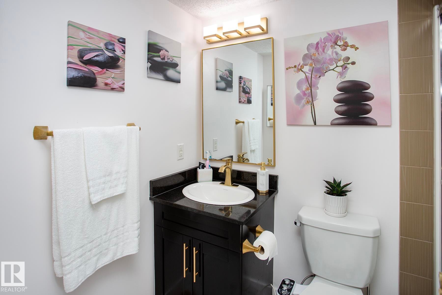 Vanity with dark countertop and gold-finish faucet, framed mirror, and coordinated wall-mounted lighting - #201 1070 Mcconachie Bv Nw, Edmonton, AB