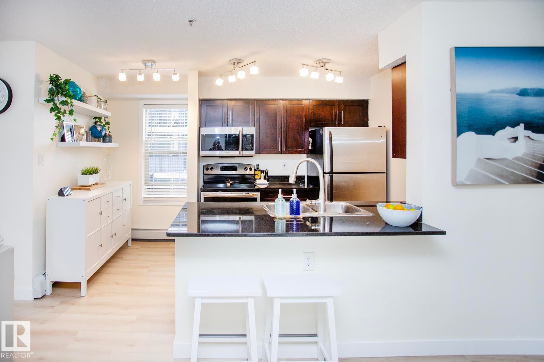 Kitchen featuring dark wood cabinetry, stainless steel appliances, a black countertop breakfast bar, and track lighting - #201 1070 Mcconachie Bv Nw, Edmonton, AB