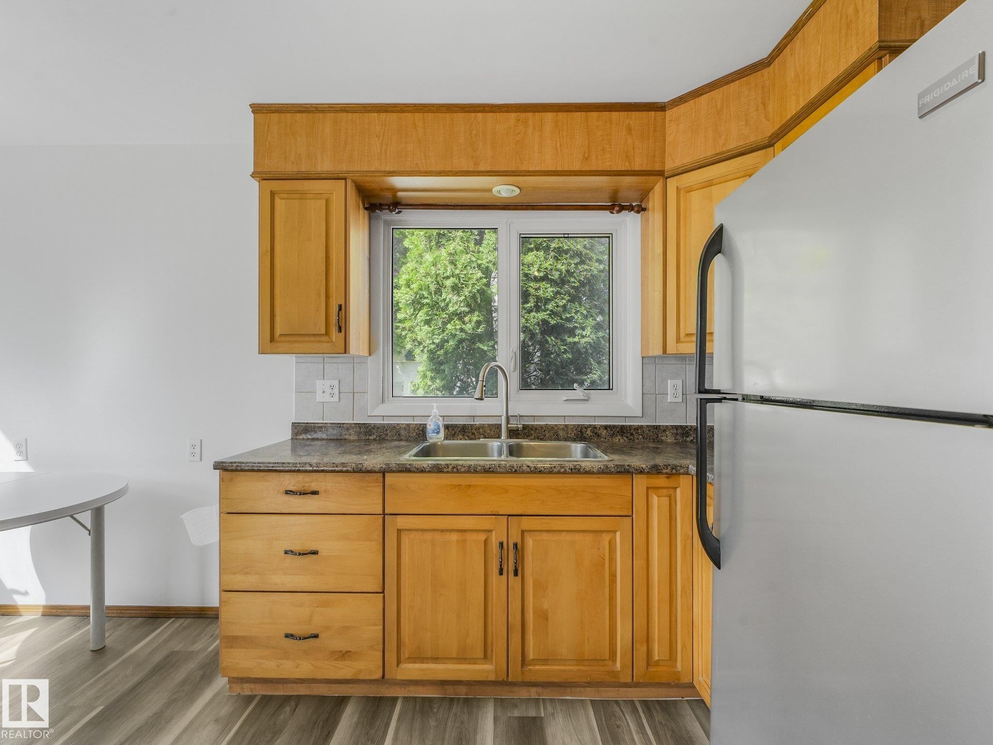 Wood-finish cabinetry, stainless steel sink, window over sink, white tile backsplash, and durable wood-finish flooring - 16428 88 Avenue Nw, Edmonton, AB