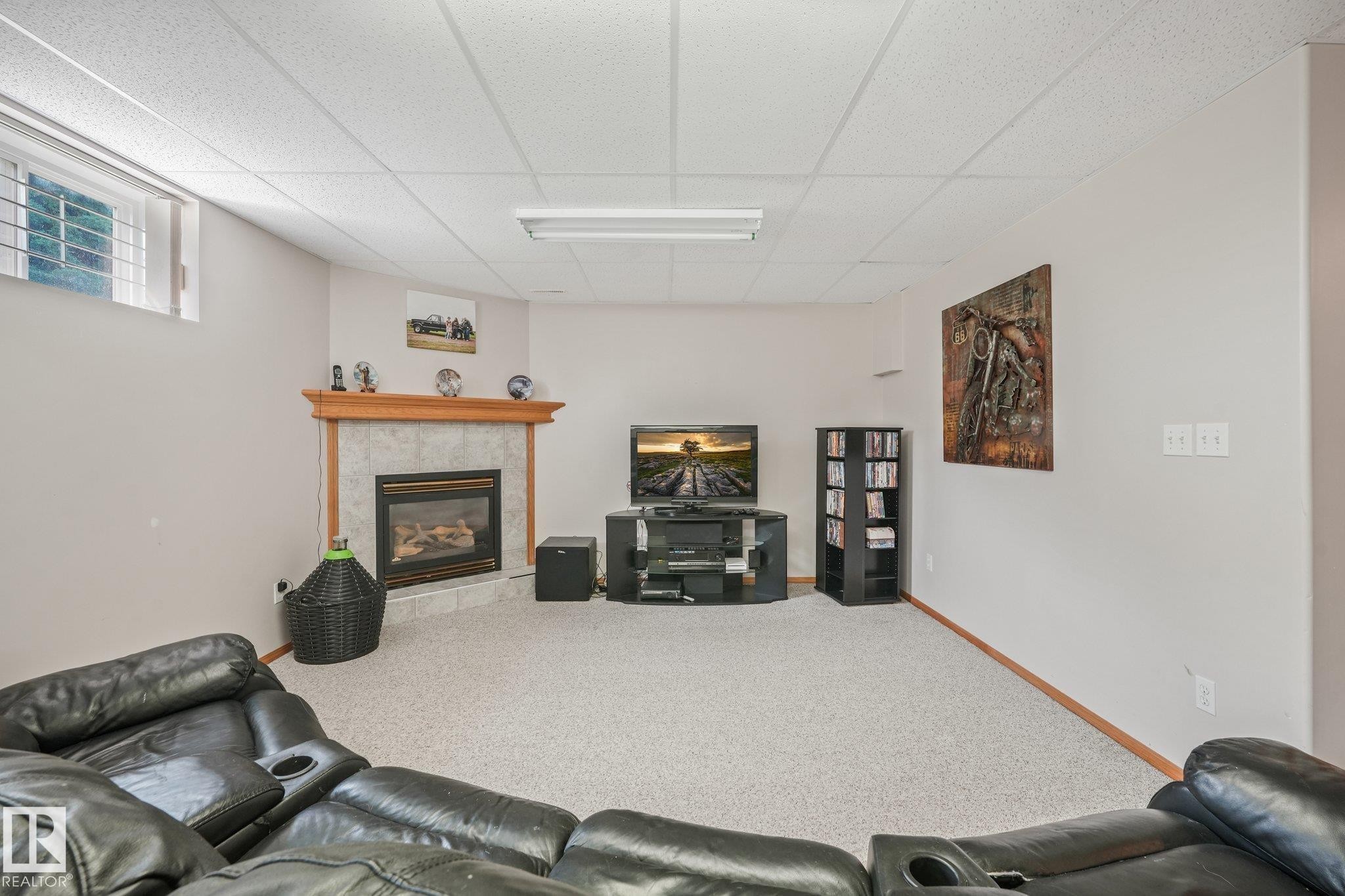 Spacious room featuring a corner fireplace with a tiled surround and wood mantel, light-colored carpet flooring, a drop ceiling with fluorescent lighting, and a high-set window - 54 Buskmose Dr, Rural Wetaskiwin County, AB - Indoor With Fireplace