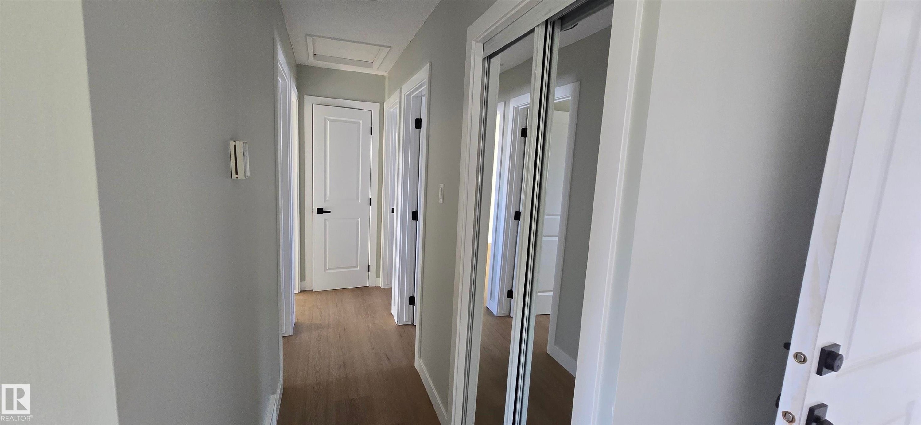 Hallway featuring wood-finish flooring, white paneled doors with matte black hardware, and mirrored closet doors - 13812 63 Street Nw, Edmonton, AB