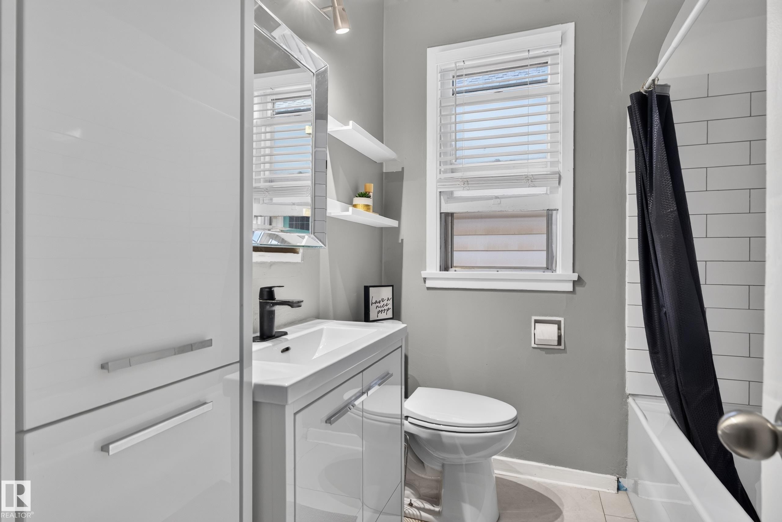 Modern white vanity with integrated sink and matte black faucet, wall-mounted mirror, white subway tile shower surround, and white window blinds - 11406 83 Street Nw, Edmonton, AB