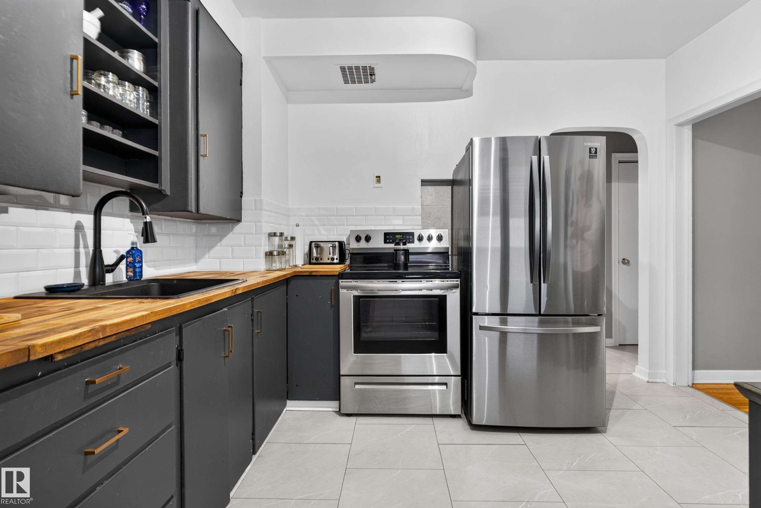 Updated kitchen featuring stainless steel appliances, dark cabinetry, wood-finish countertops, subway tile backsplash, and light tile flooring - 11406 83 Street Nw, Edmonton, AB