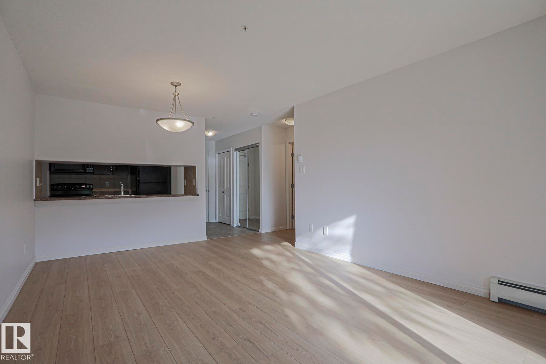 Open-concept living area featuring wood-finish flooring, a contemporary pendant light fixture, and a pass-through window to the kitchen with dark countertops - #108 1204 156 St Nw, Edmonton, AB