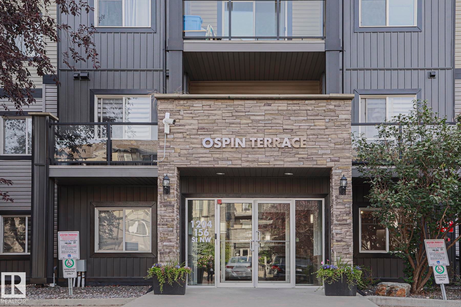 Stone-clad entrance with glass double doors, featuring integrated exterior lighting and a building nameplate - #108 1204 156 St Nw, Edmonton, AB