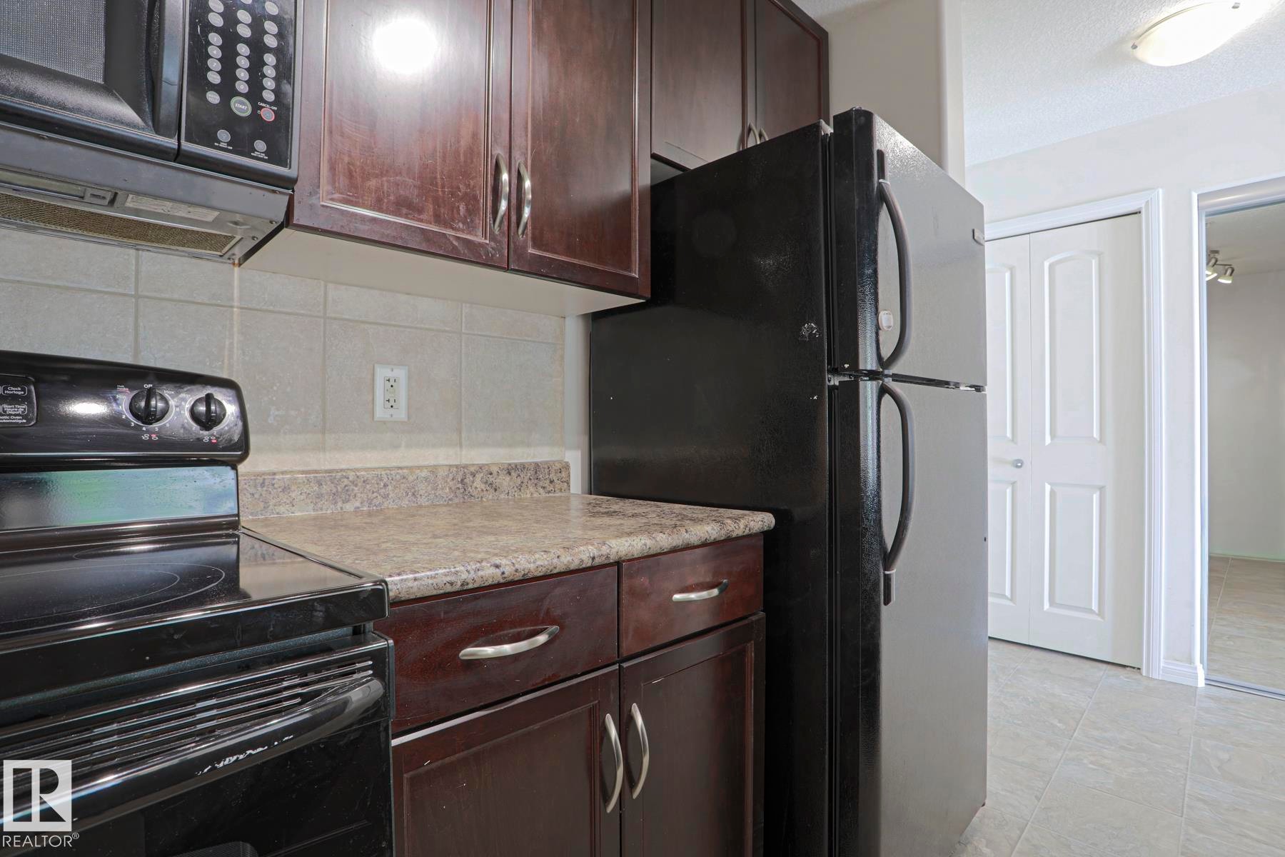 Kitchen featuring dark wood-finish cabinetry, light-toned laminate countertops, and a full suite of black appliances, including a refrigerator, range, and microwave - #108 1204 156 St Nw, Edmonton, AB