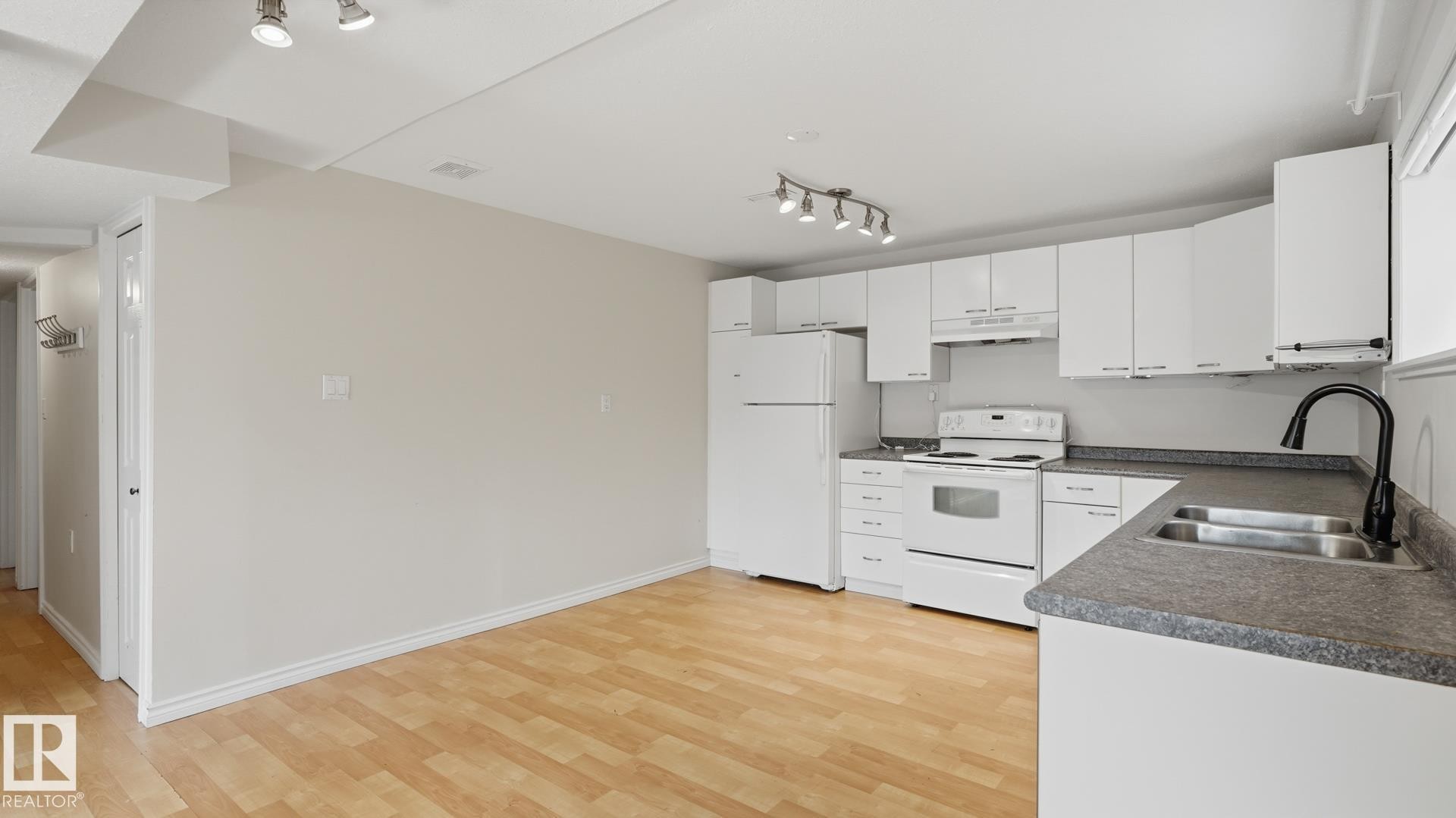 Open-concept living space with wood-finish flooring, a kitchen featuring white cabinetry, a dual basin stainless steel sink with a matte black faucet, and track lighting - 12208 146 Avenue Nw, Edmonton, AB