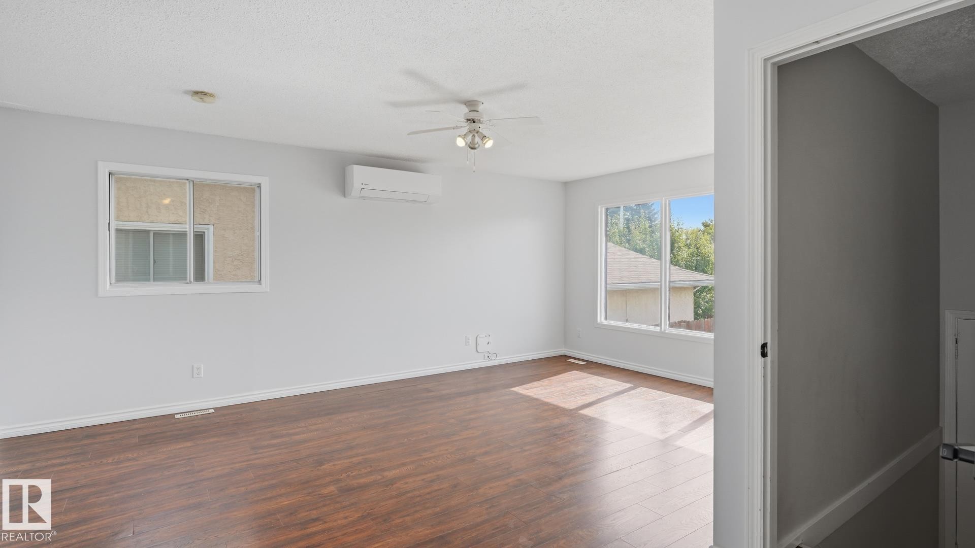 Spacious room featuring wood-finish flooring, multiple windows, a ceiling fan, and a wall-mounted climate control unit - 12208 146 Avenue Nw, Edmonton, AB